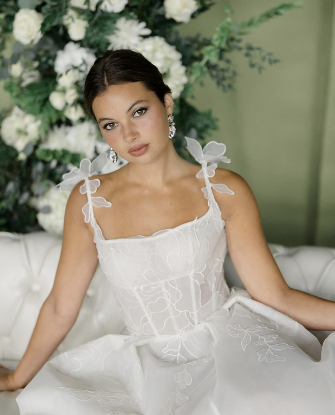 Young woman in a white dress with bows on shoulders, sitting on a white couch with floral background.