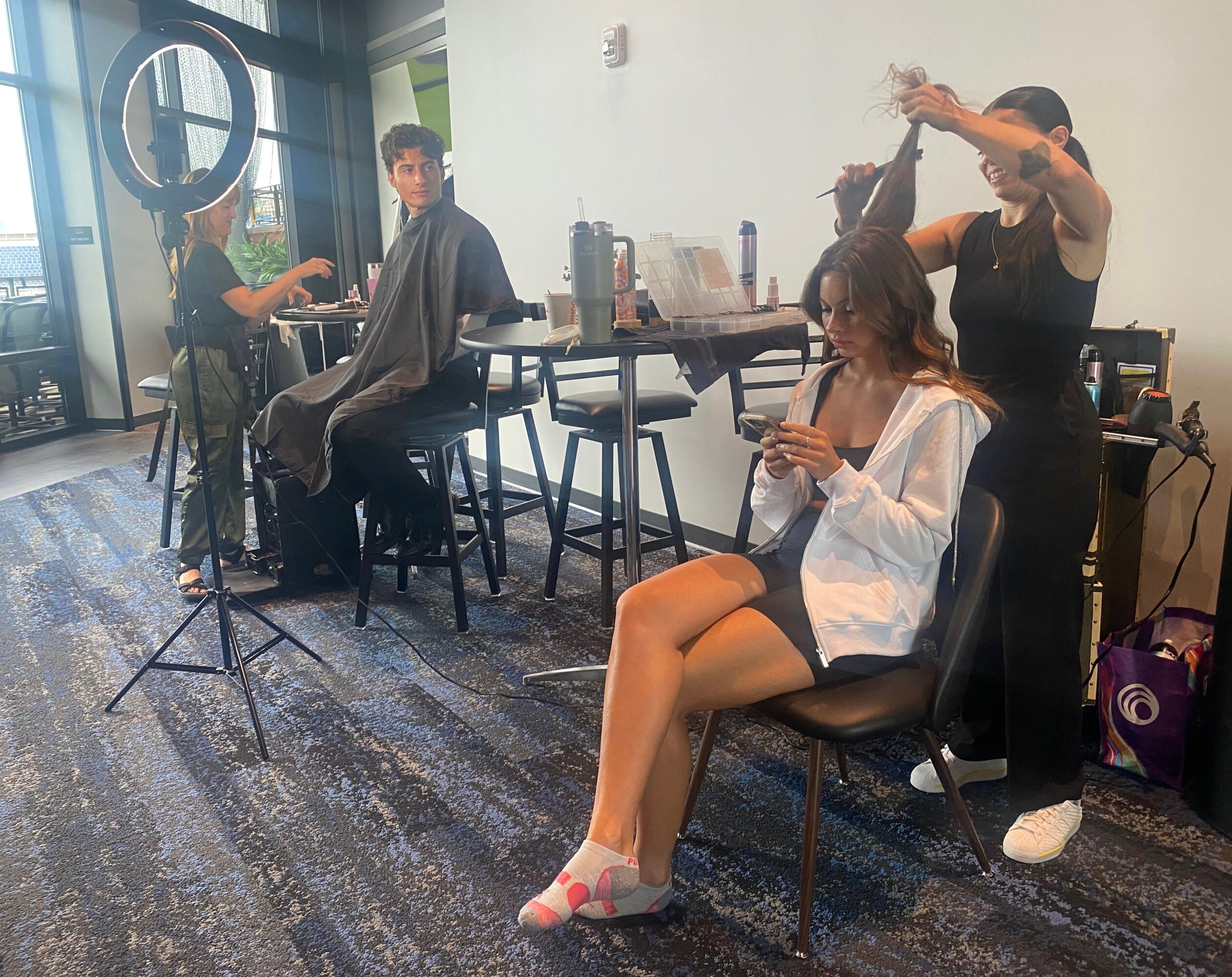A woman sits on a chair using her phone, with two hairstylists working on her hair in a salon.