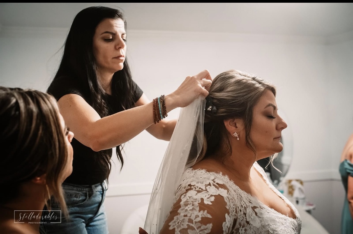 A woman with long dark hair styles a bride's hair, who is wearing a lace wedding dress and veil, in a room.
