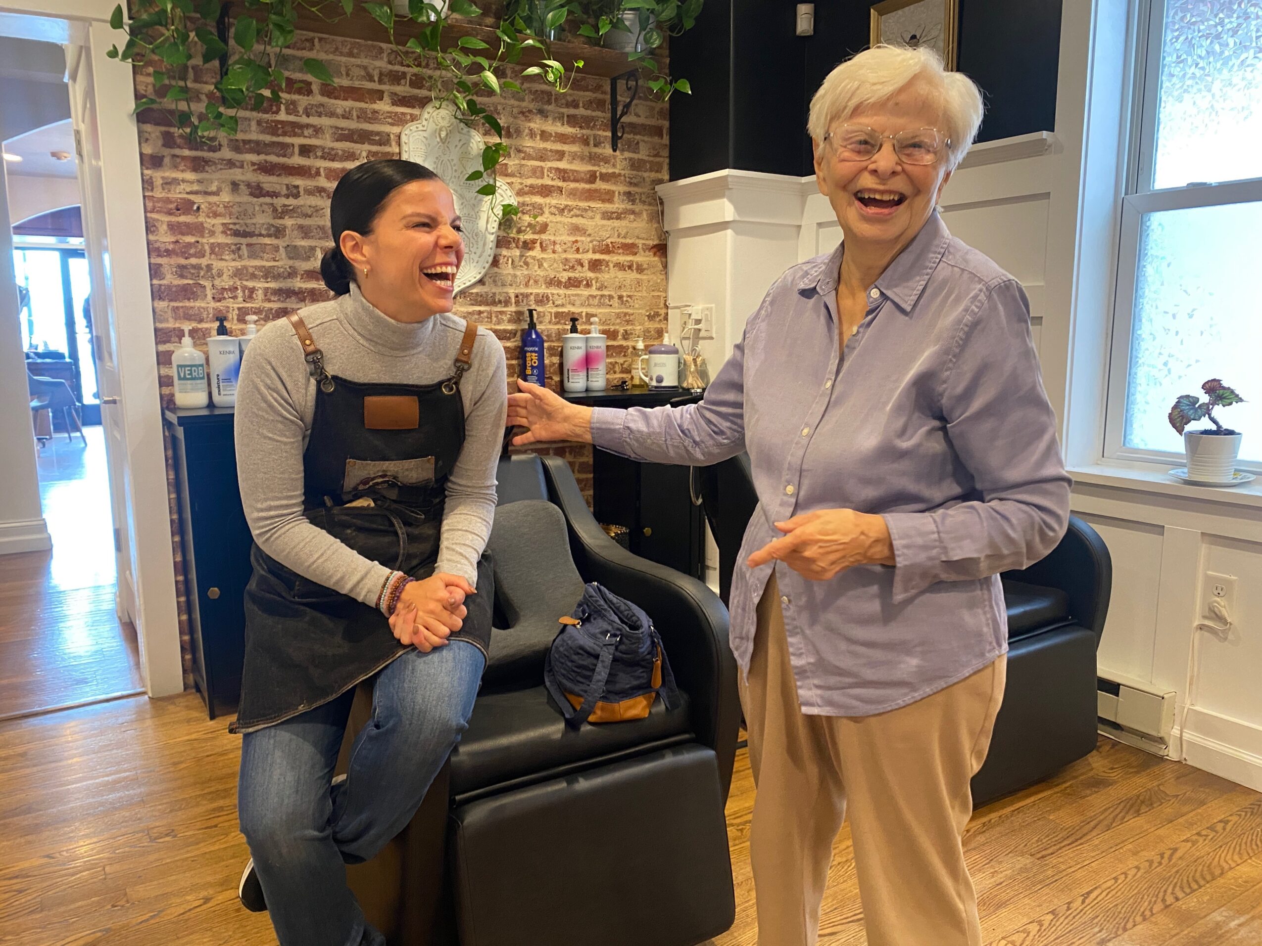 Two women laughing and talking in a cozy room with brick wall and wooden floor.