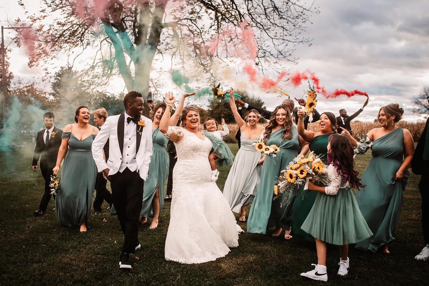 Group of people celebrating outdoors, some holding colorful smoke flares, with a bride in a white dress and others in teal dresses.