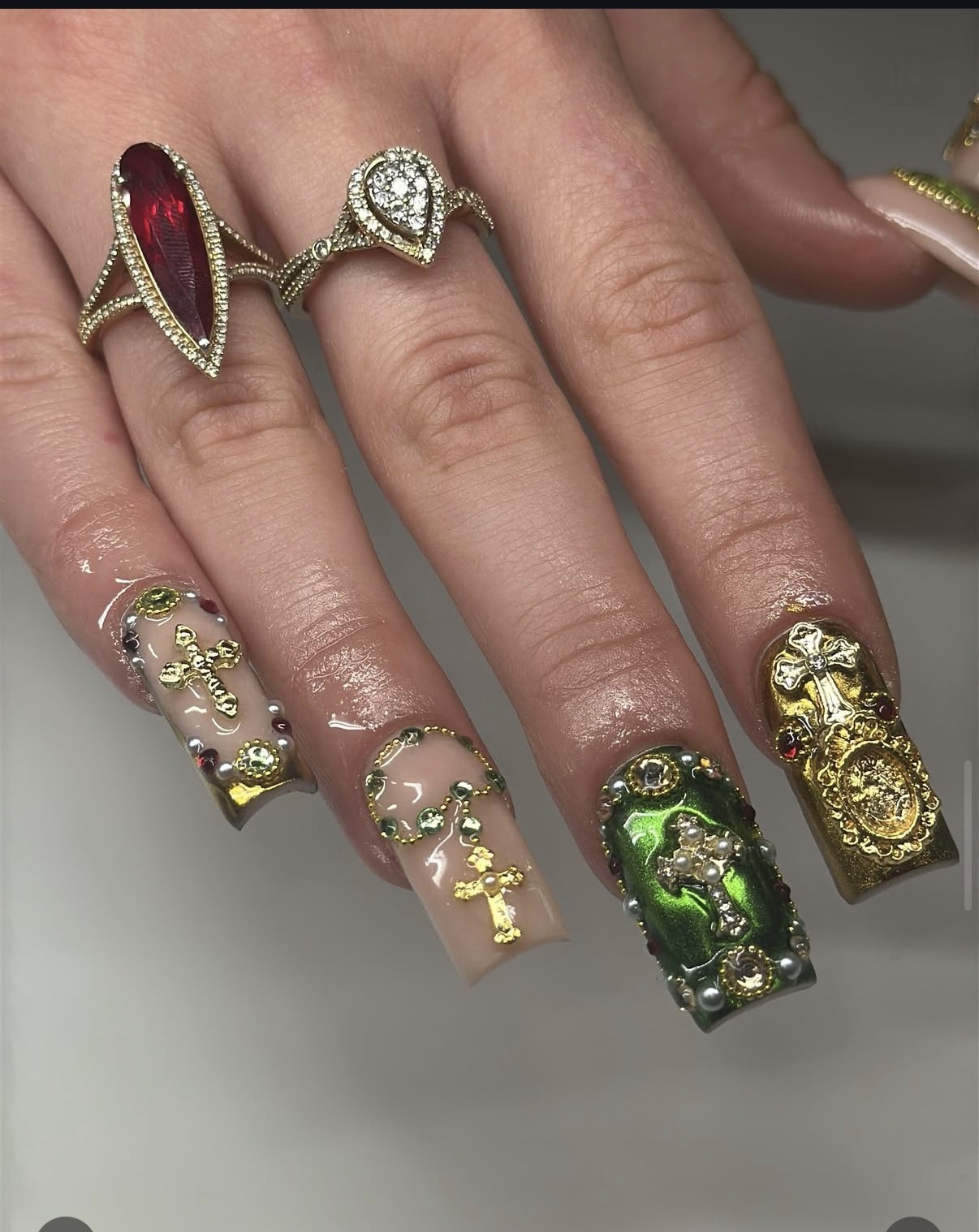 Hand with decorated nails and multiple rings, including a large red and gold ring, and a ring with a diamond. Nail art features gold, green, and black designs.
