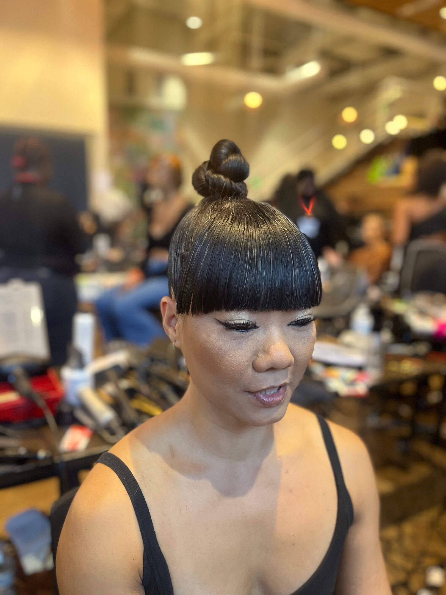 Woman with a high bun hairstyle and straight bangs, wearing a black spaghetti strap top, in a busy indoor setting.