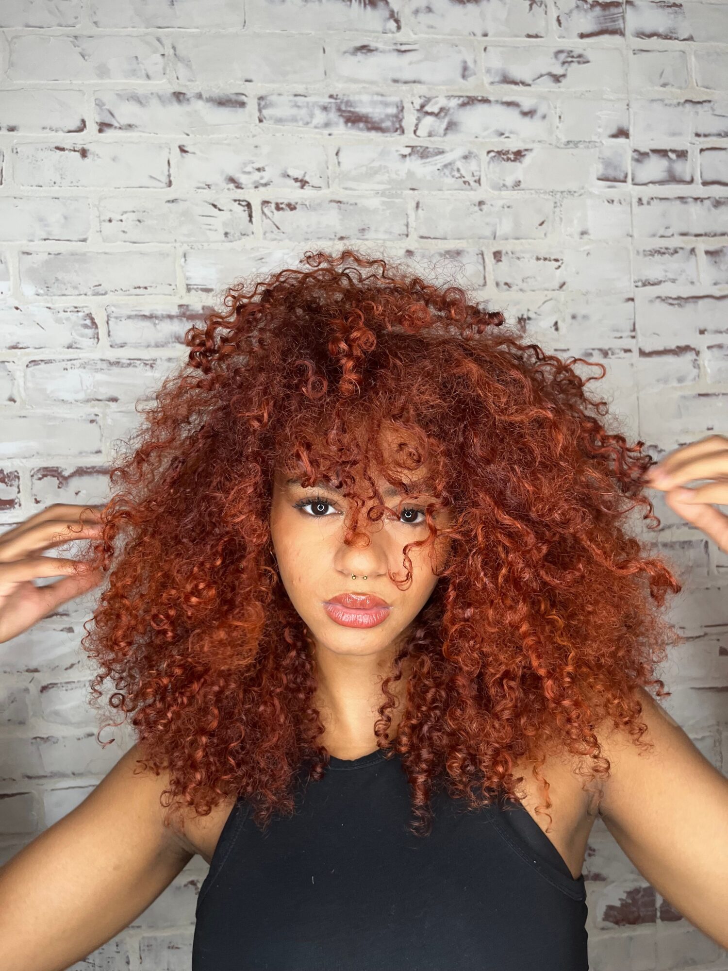Woman with curly red hair lying on a white brick wall, wearing a black top, looking at the camera.