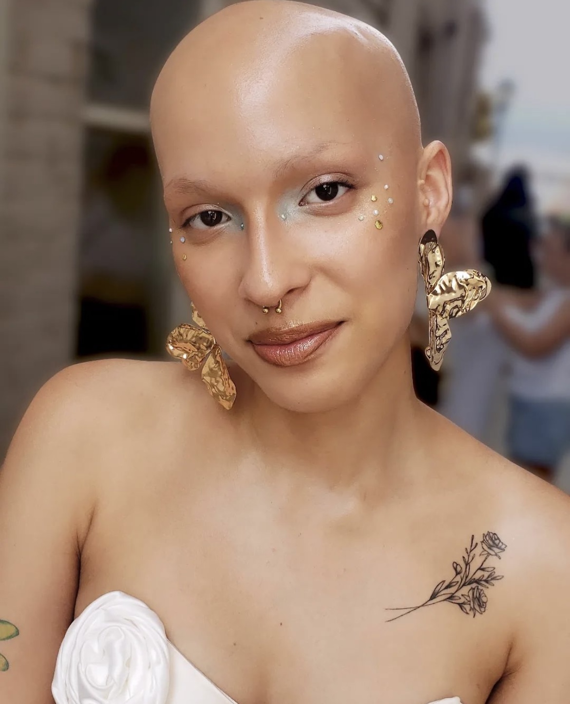 Bald person with large gold earrings, nose piercing, and a tattoo on shoulder, smiling outdoors.