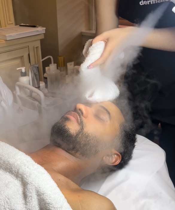 Person receiving a facial treatment with steam and a practitioner applying a mask or cream, lying on a bed.