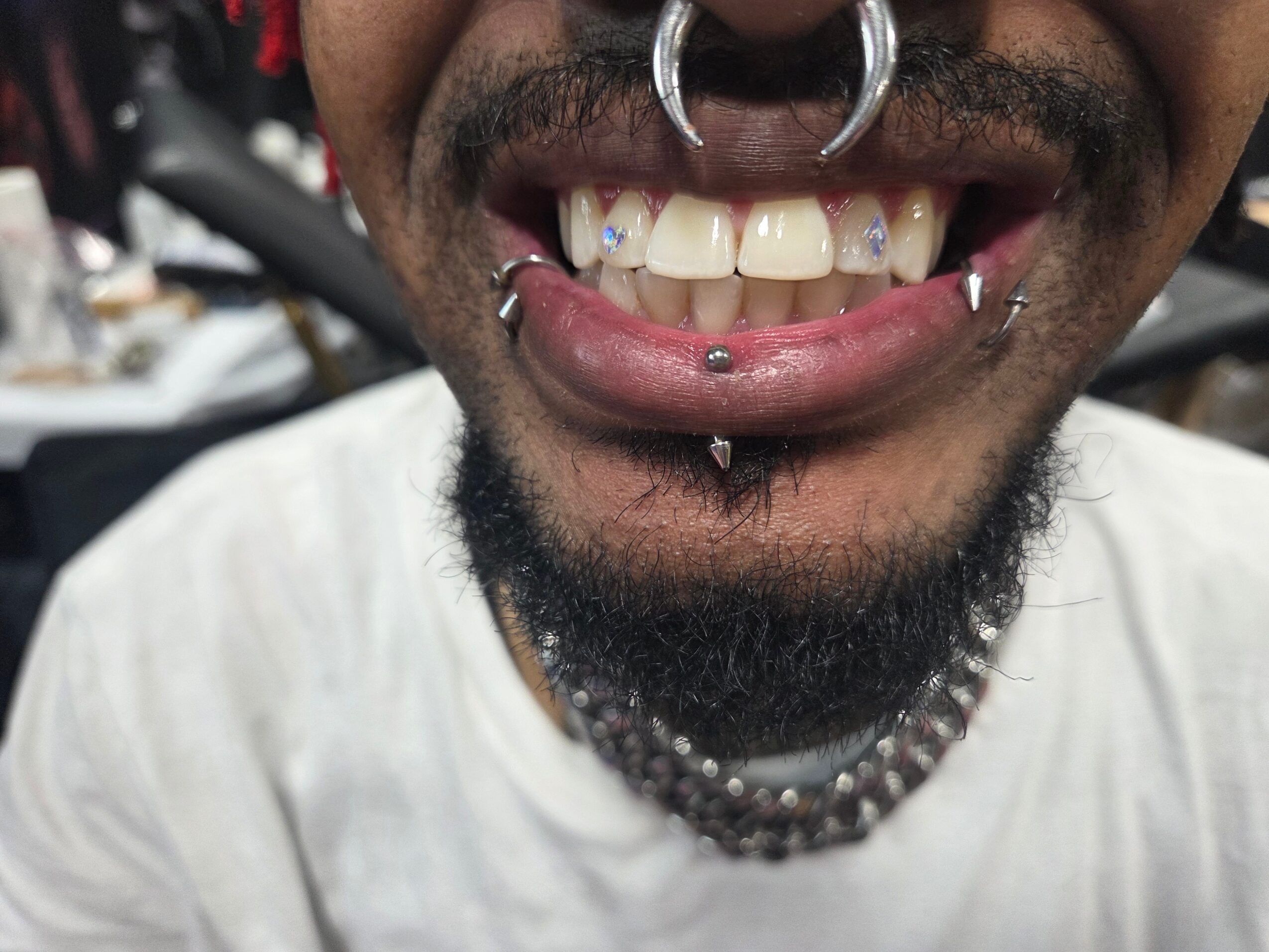 Close-up of a person's mouth showing multiple facial piercings, teeth, and a beard, with a blurred background.
