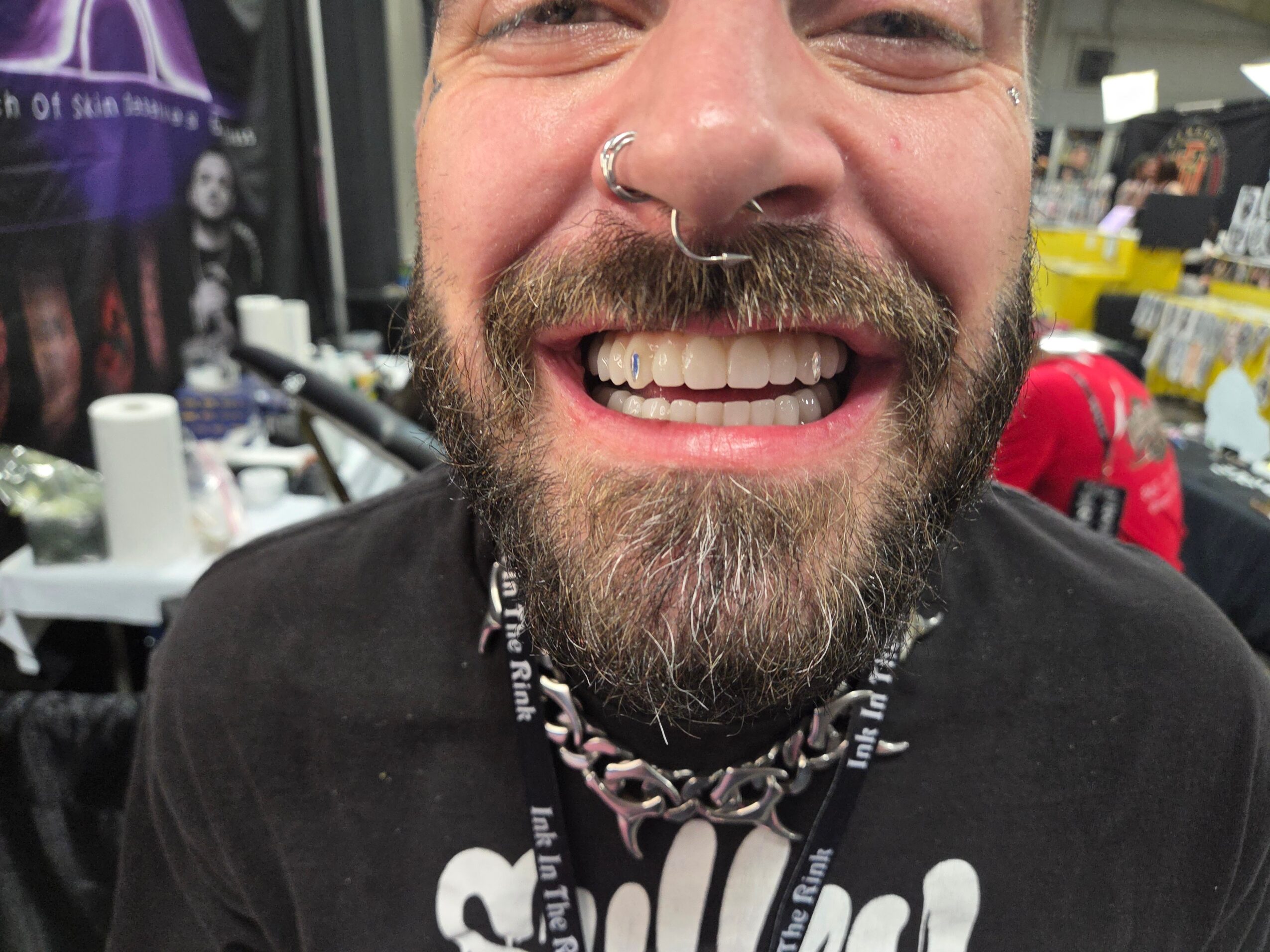 Close-up of smiling person with a beard, nose ring, and layered necklaces, in a busy indoor setting.