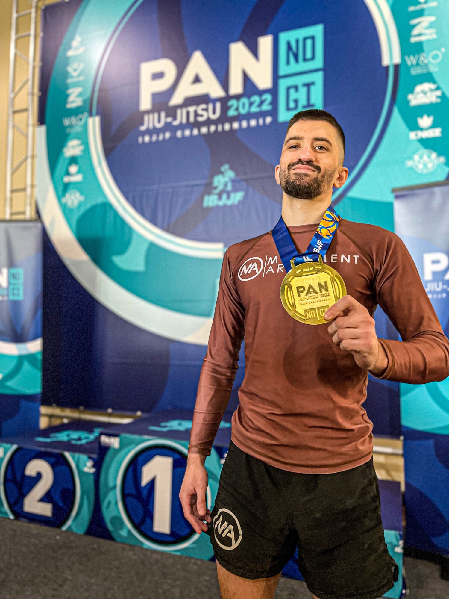 Man holding a gold medal at a sports event, standing in front of a large circular banner with text and logos.