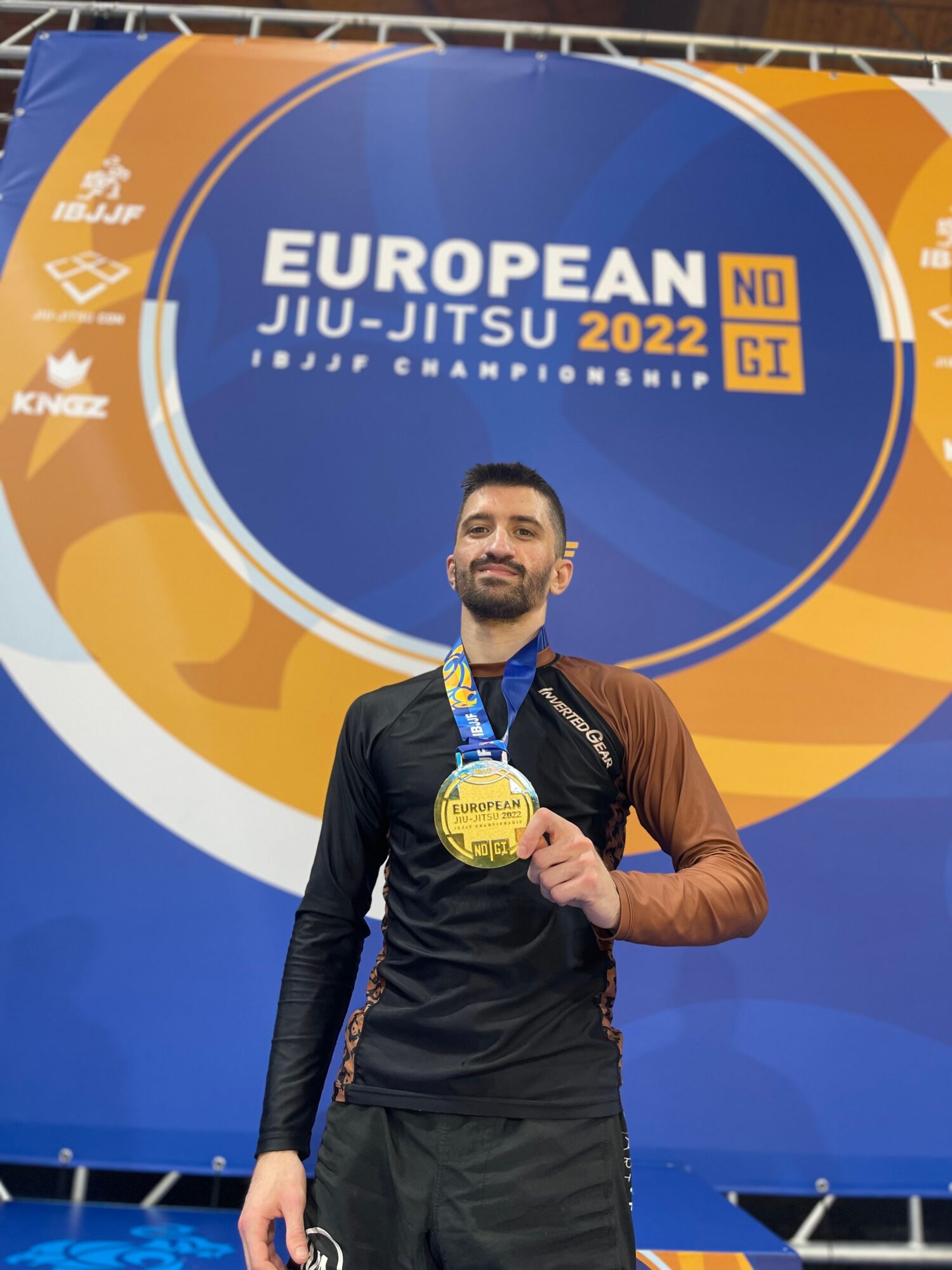 Male athlete holding a medal at European Jiu-Jitsu Championship 2022, standing in front of event banner.