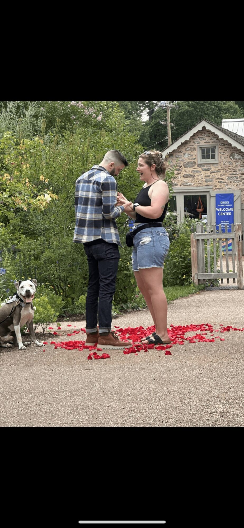 Two people stand on a path with a dog sitting nearby, surrounded by greenery and a building in the background.