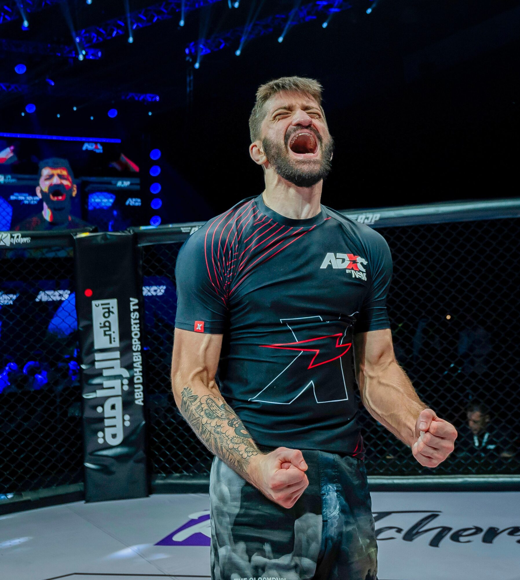 Mixed martial artist inside the cage, shouting with clenched fists, wearing a black and red shirt, tattoos on arm, cheering.