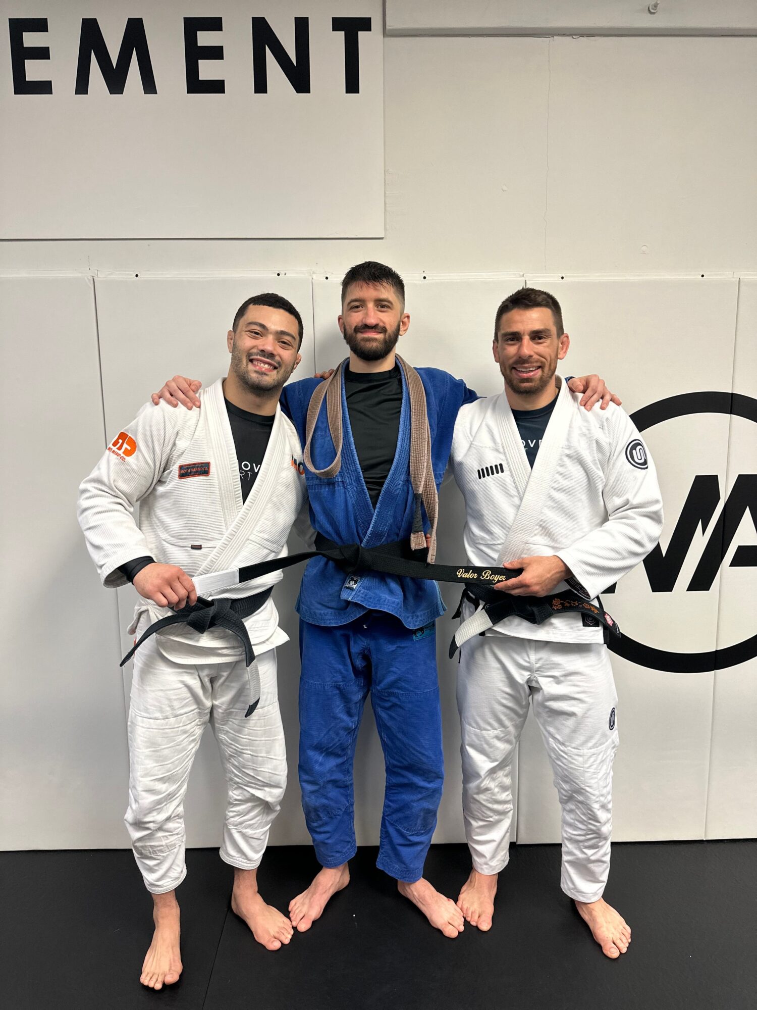 Three people in martial arts uniforms with black belts, standing on a black mat, smiling, with a white wall and logo behind them.