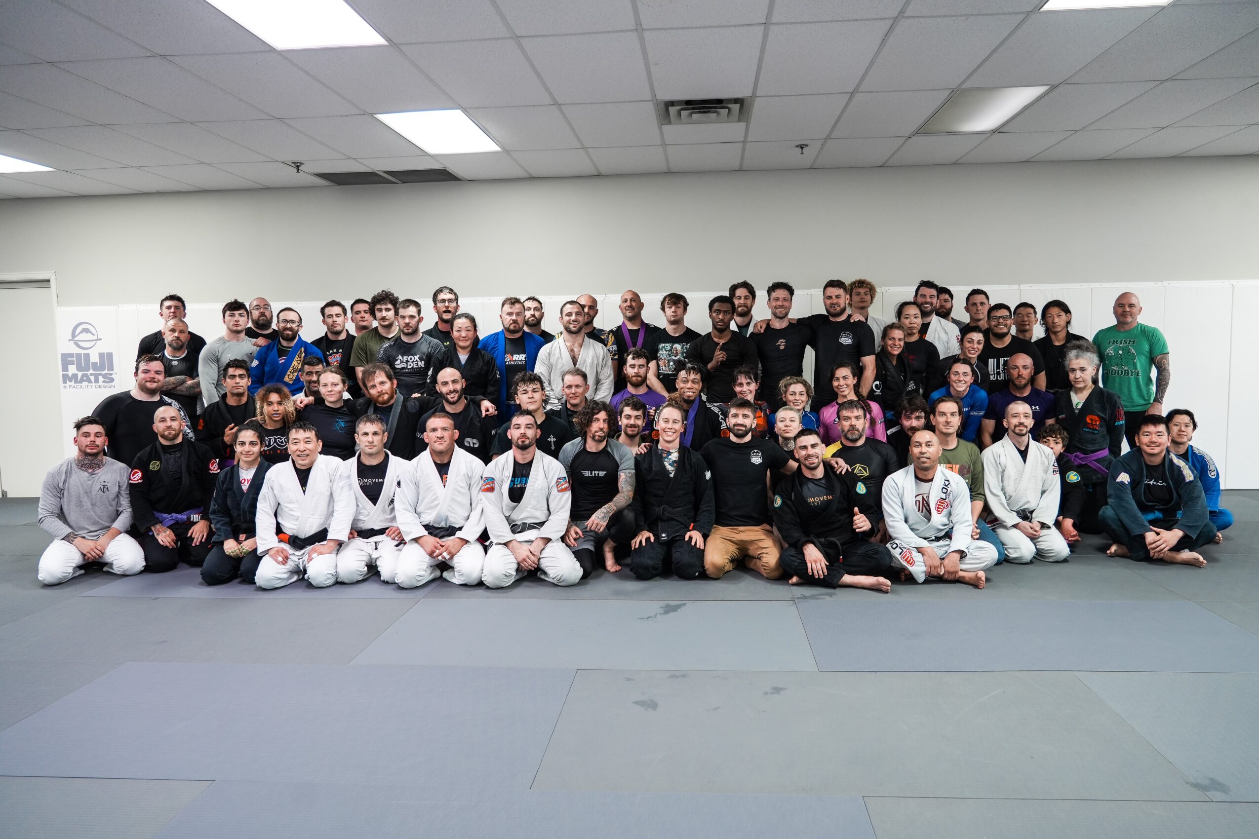 Group of people in martial arts uniforms and casual clothing posing in a dojo, some kneeling, some standing, with a white wall background.