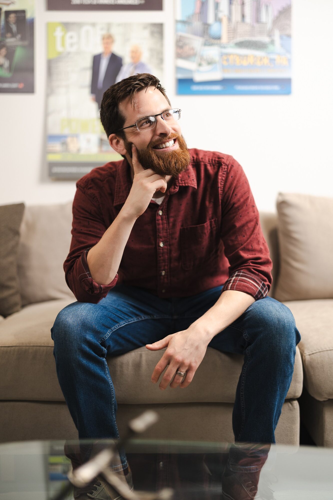 Man with glasses and a beard, smiling, sitting on a sofa in a room with posters on the wall behind him.
