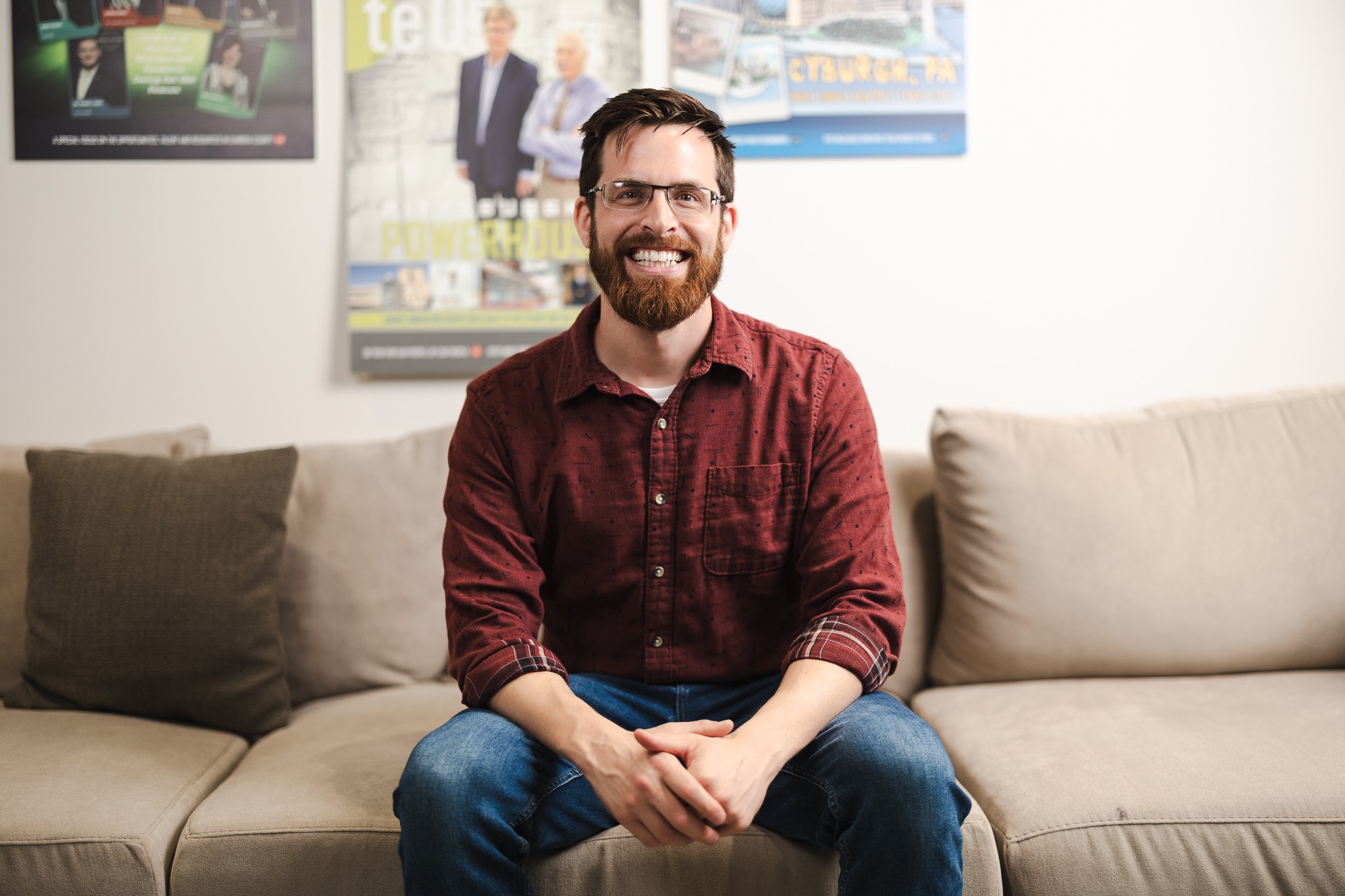 Smiling man with glasses and beard sitting on a beige sofa in a room with posters on the wall.