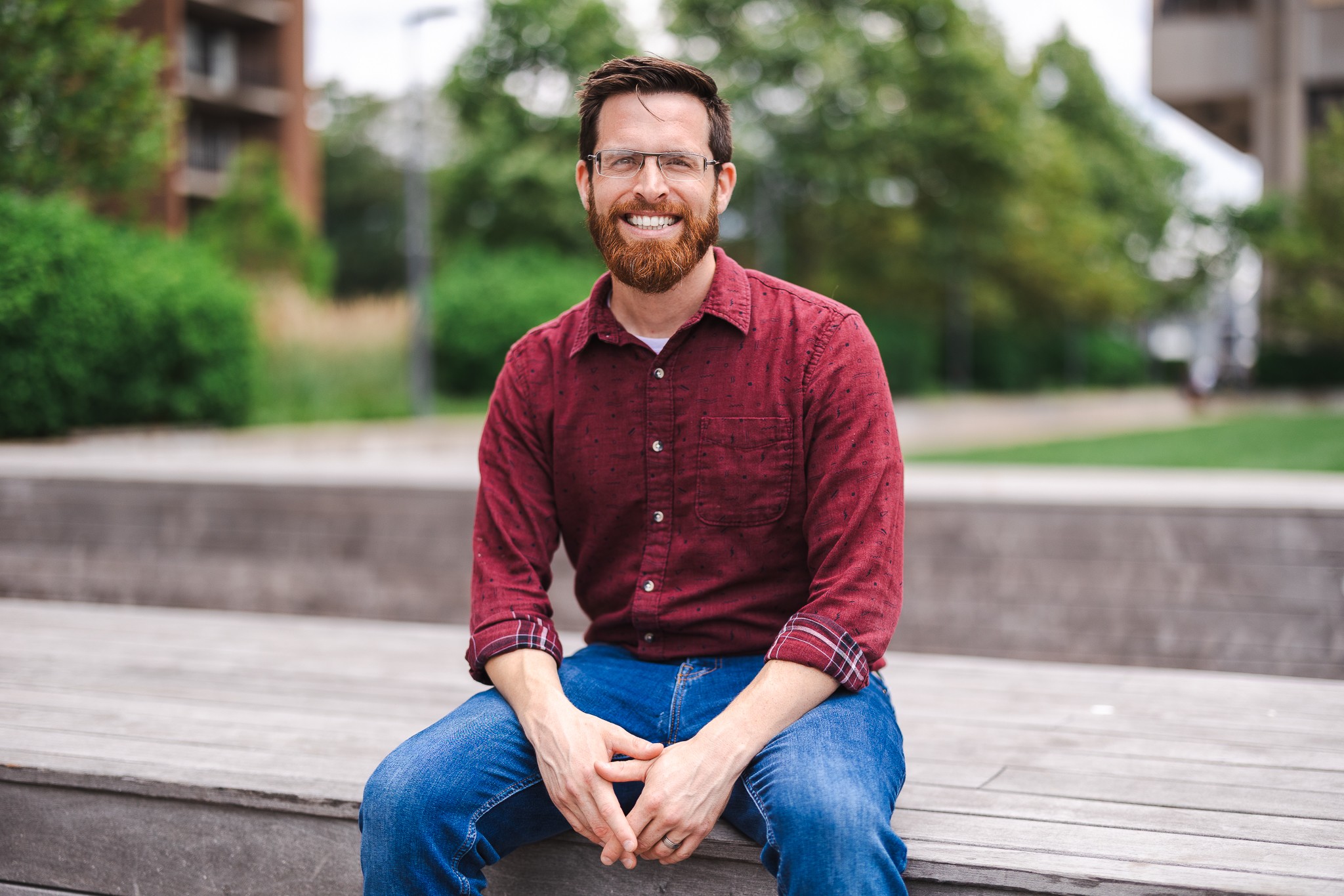 Man with glasses and beard sitting outdoors on a bench, smiling, wearing a red shirt and blue jeans.