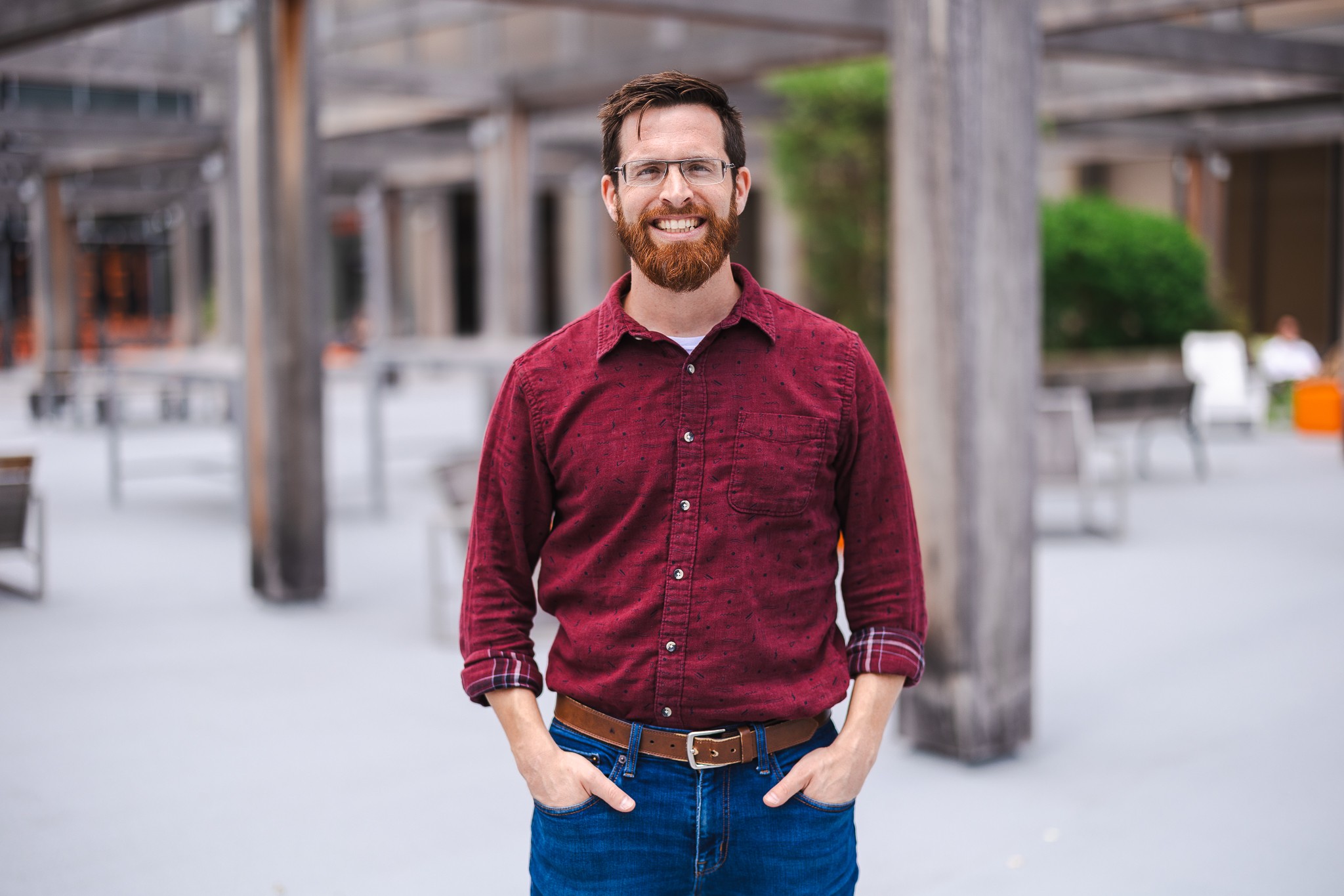 Man with glasses and beard wearing a red shirt, smiling outdoors with wooden structures in background.