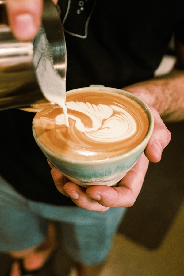 Person pouring milk into a cup of coffee with latte art, held in hand, background blurred.