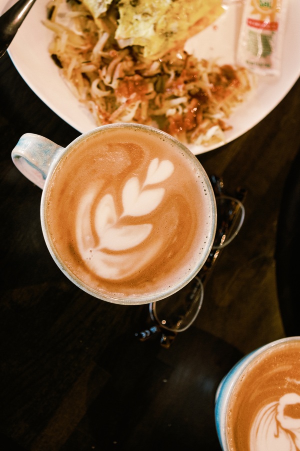 A glass mug of hot chocolate with latte art on top, viewed from above, with a plate of food in the background.
