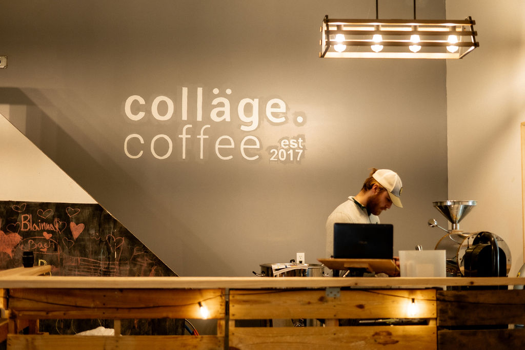 Person working behind a coffee counter with a sign reading 'collage coffee est 2017' on the wall, modern lighting, and a chalkboard.