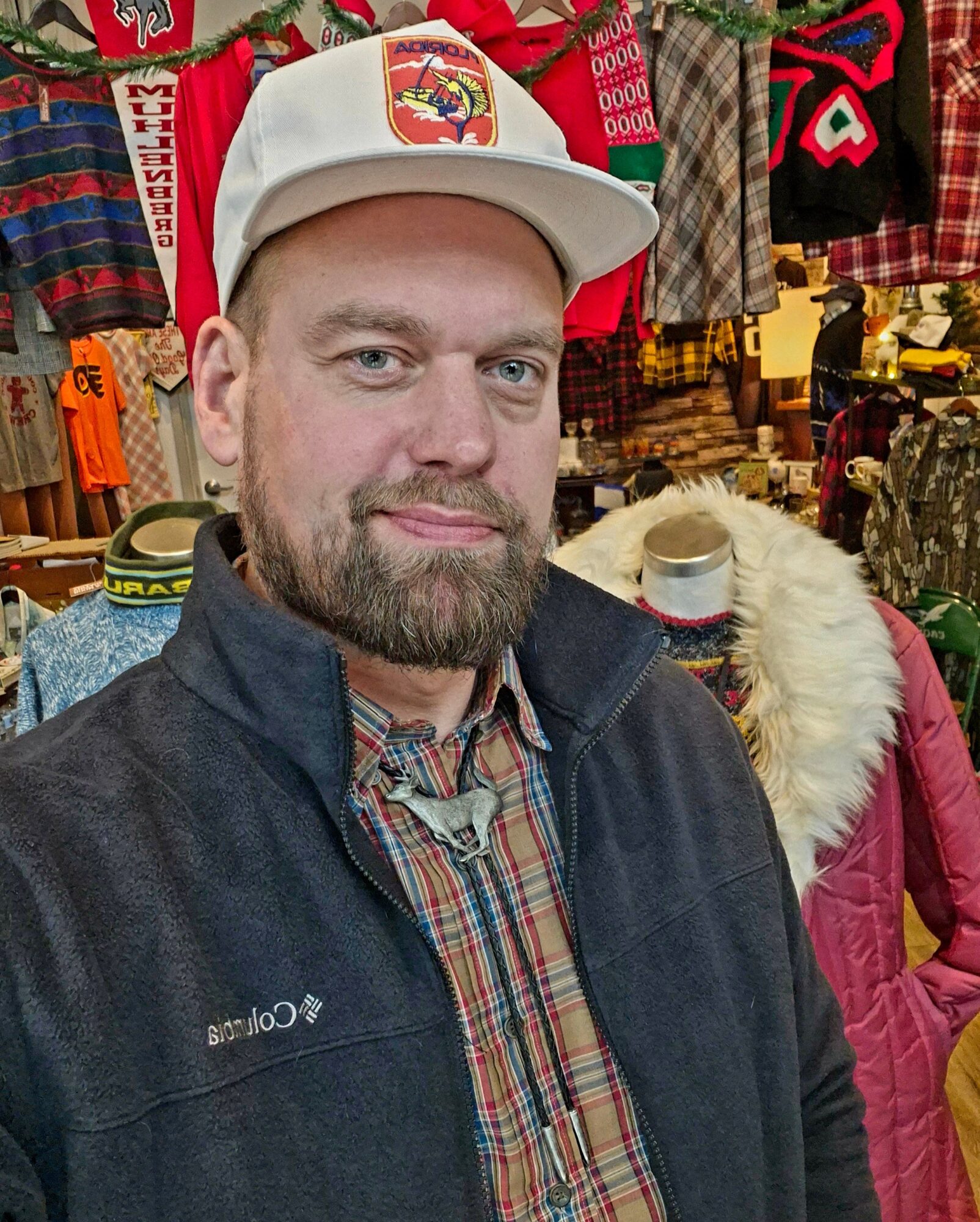 Man with a beard wearing a white cap and a dark jacket, standing in a clothing store with colorful garments around him.