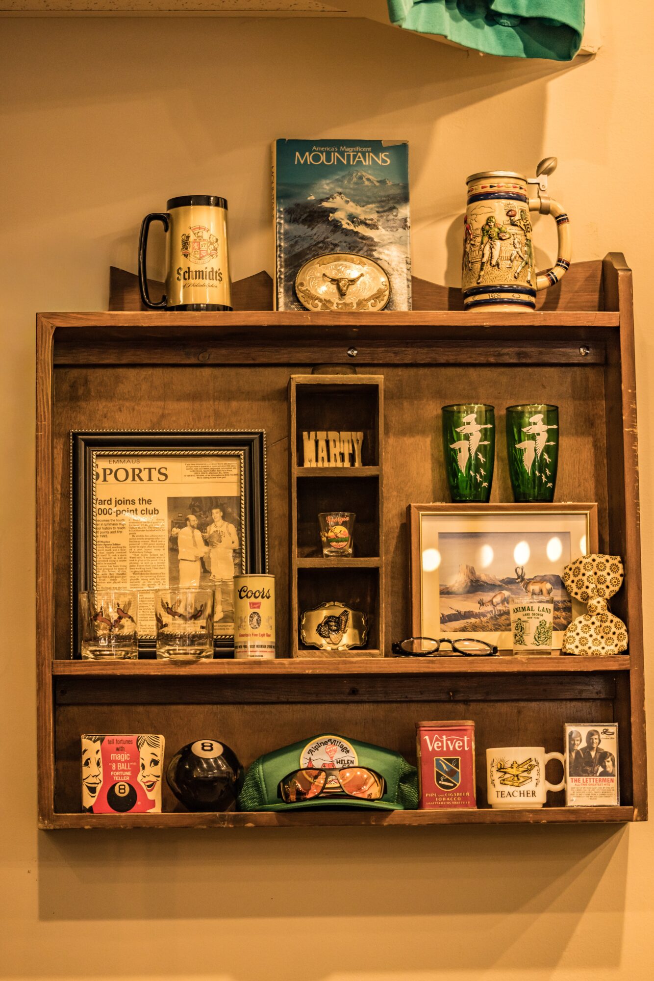 Wooden shelf with framed photos, books, and decorative items, including mugs, a clock, and small collectibles, against a beige wall.
