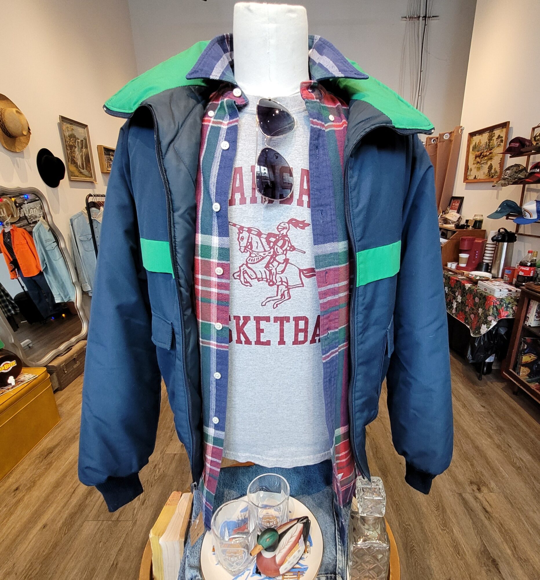 Mannequin dressed in a layered outfit with a jacket, plaid shirt, and graphic t-shirt, inside a store with various items on shelves.