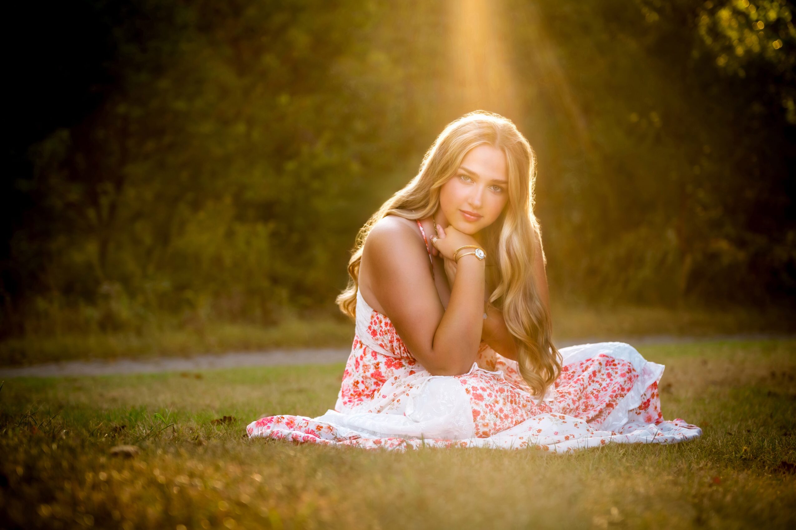 Young woman with long hair sitting on grass in a floral dress, sunlight shining down, surrounded by trees.