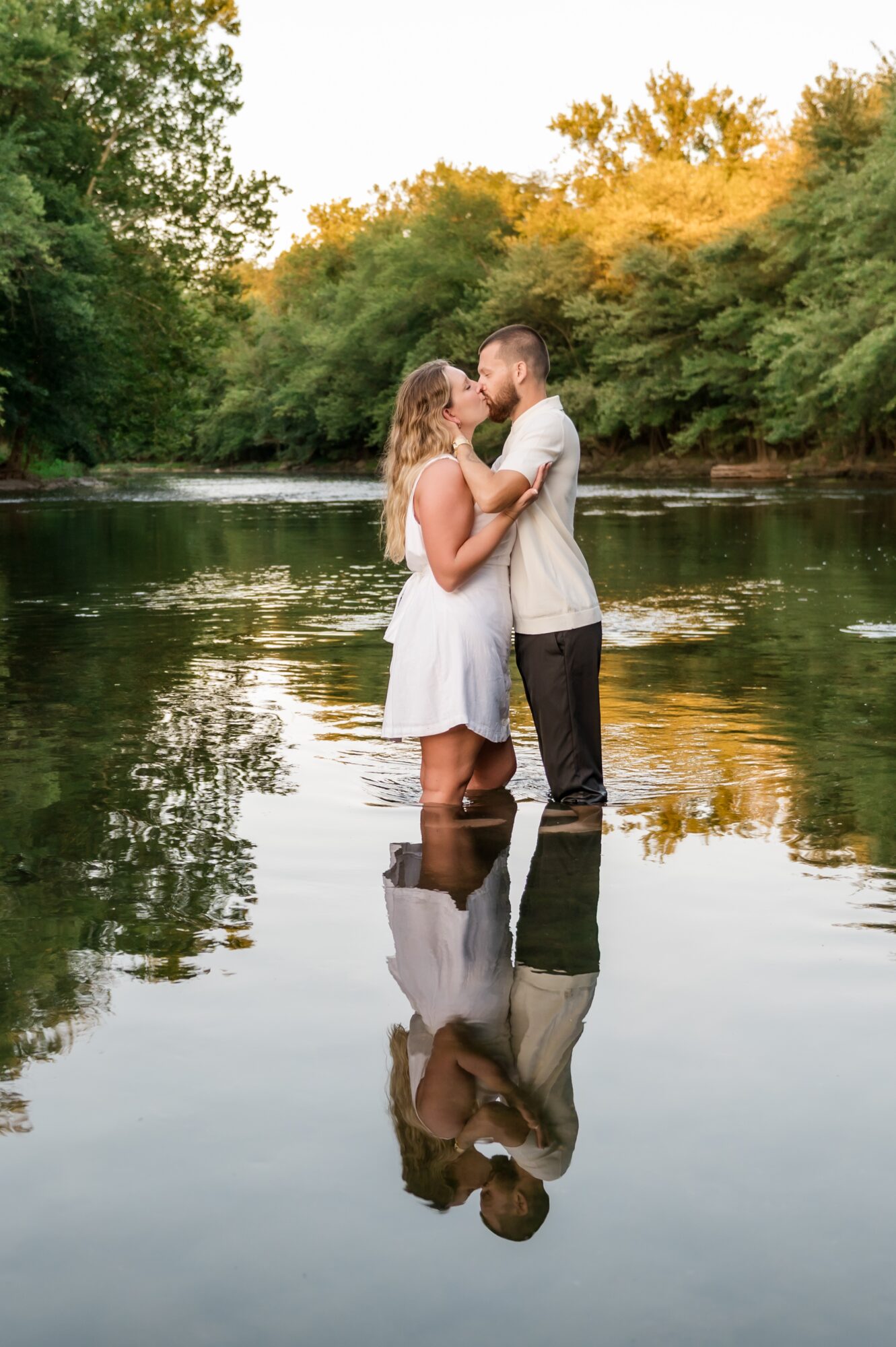 A couple stands in a river, embracing and kissing, with trees and sunlight in the background, reflected in the water.