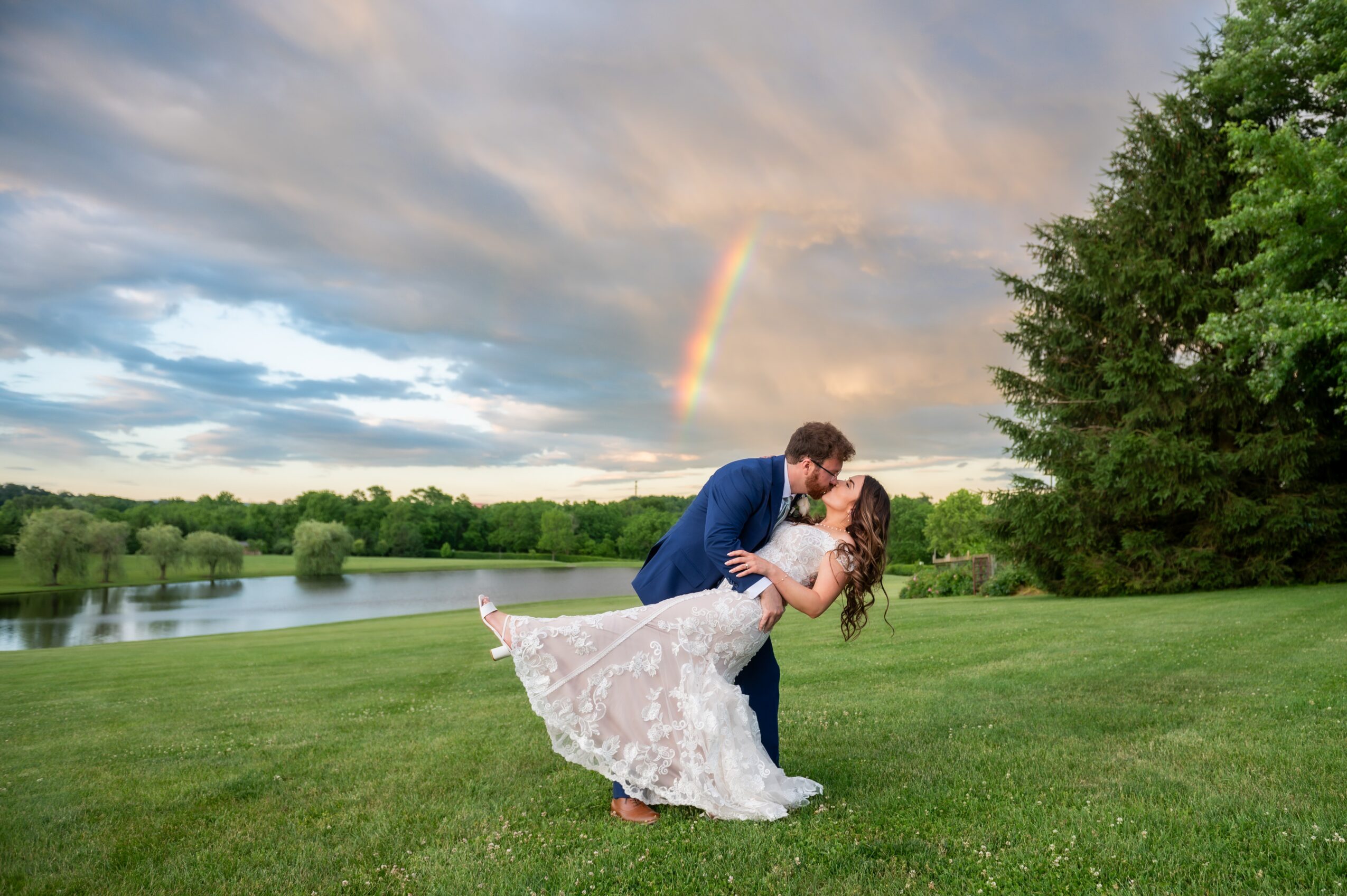 A couple dancing outdoors on a grassy field with a rainbow in the sky, trees, and a river in the background.