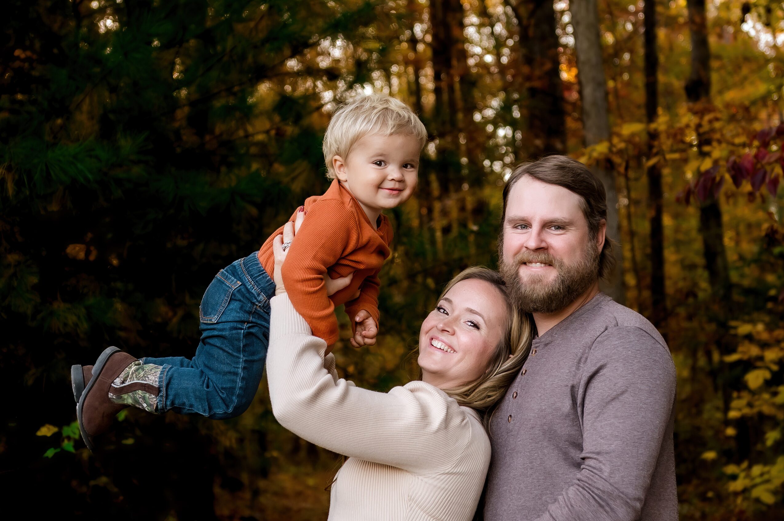 Family of three outdoors in autumn, woman lifting child, man standing beside them, trees with fall foliage in background.