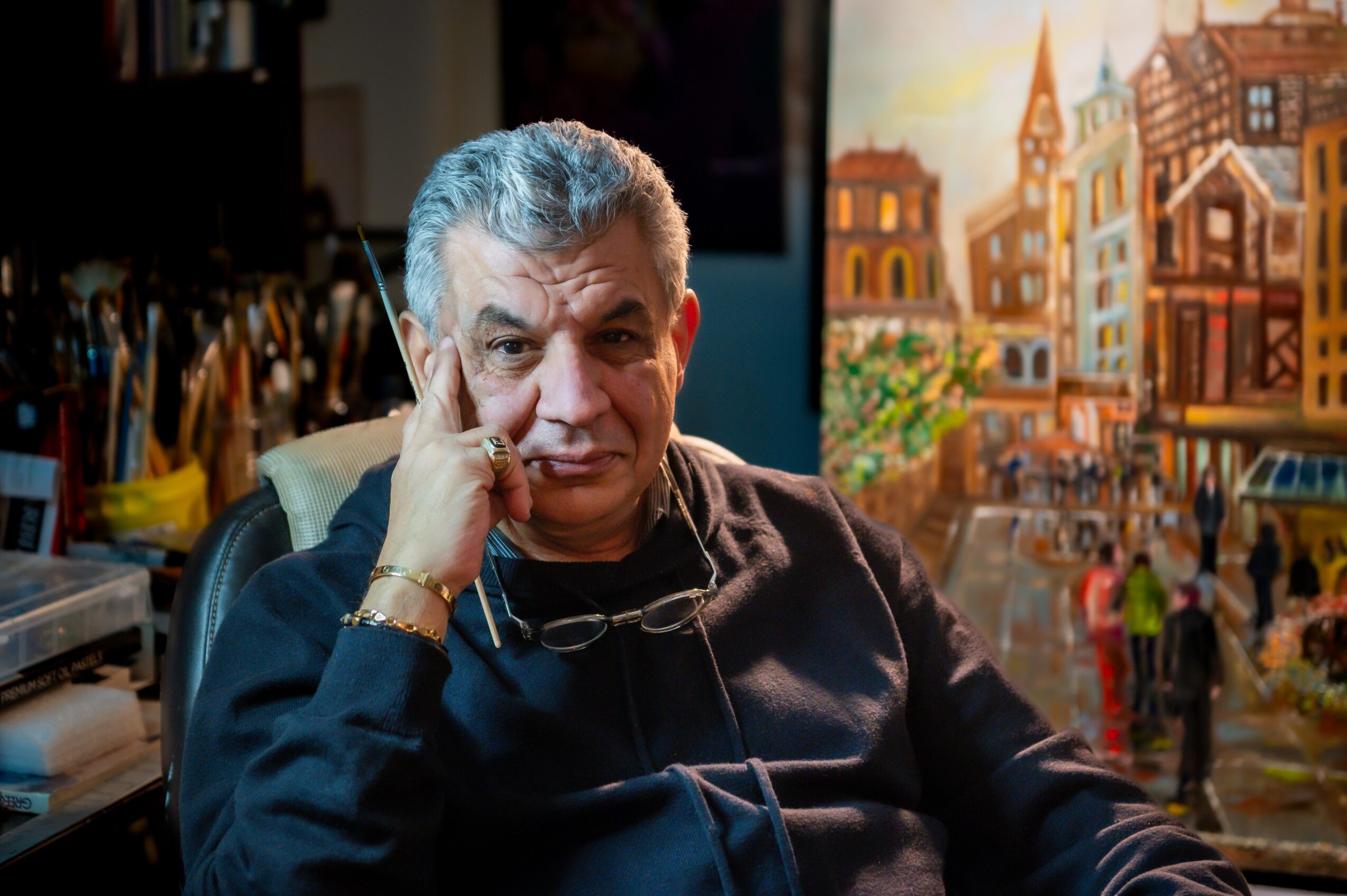 Older man with gray hair sitting indoors, resting his head on his hand, with a colorful cityscape painting in the background.