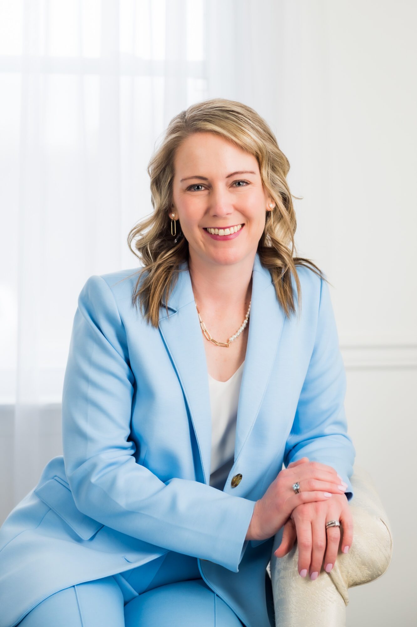 Woman with blonde hair smiling, wearing a light blue blazer and white top, sitting indoors near a window.
