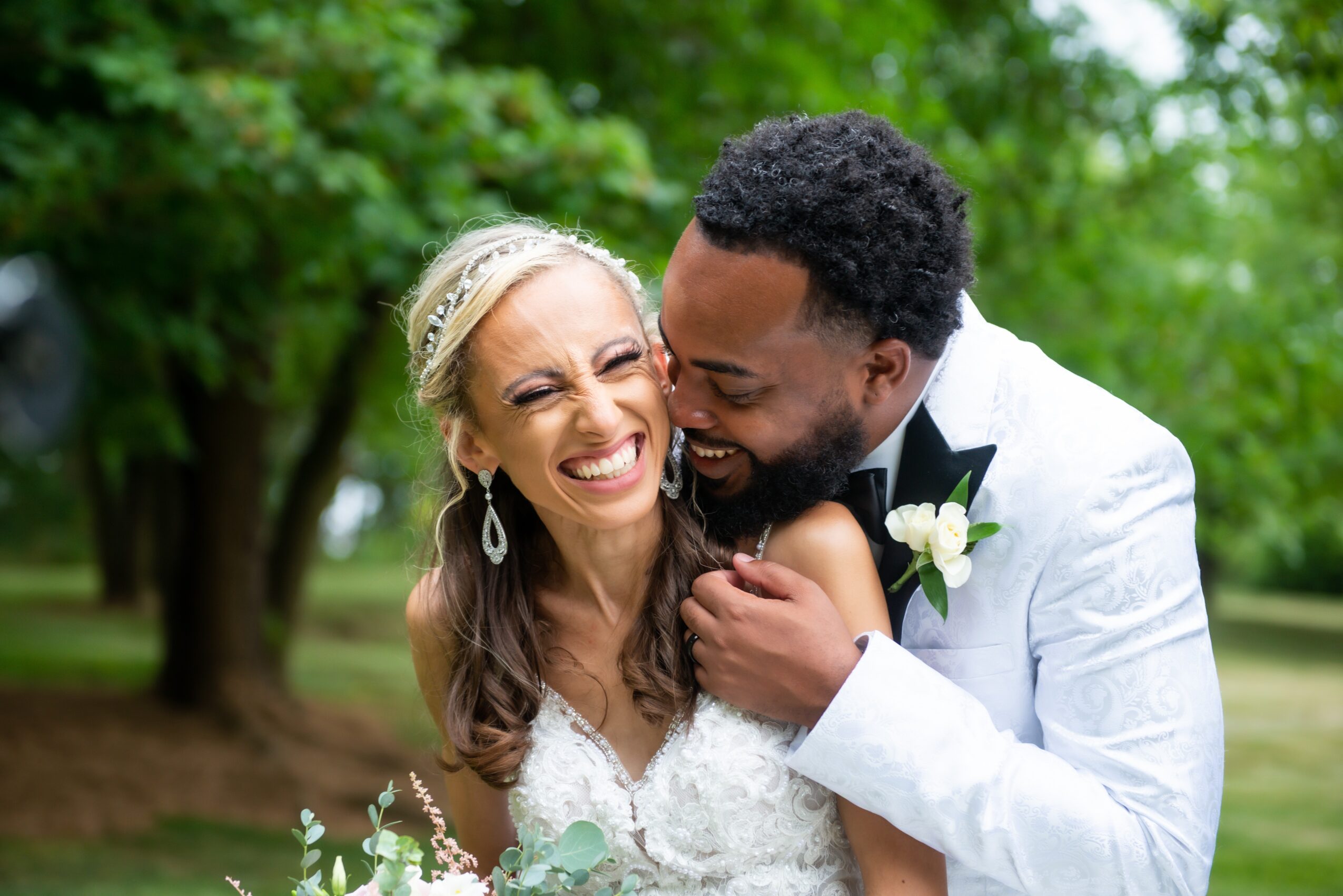 A smiling bride and groom holding a baby outdoors with green trees in the background.