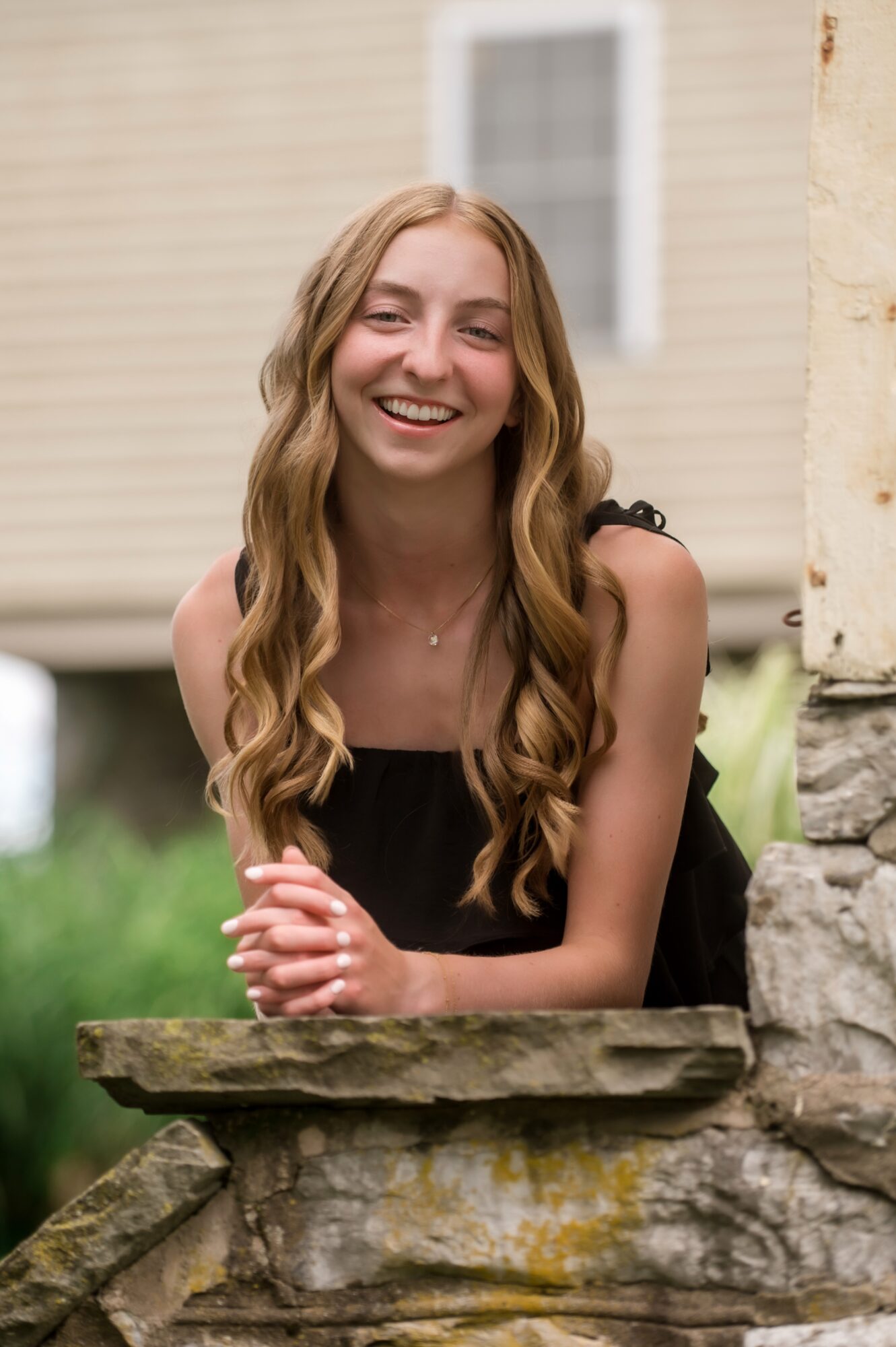 Young woman with long wavy hair smiling, leaning on a stone ledge outdoors, wearing a black top.