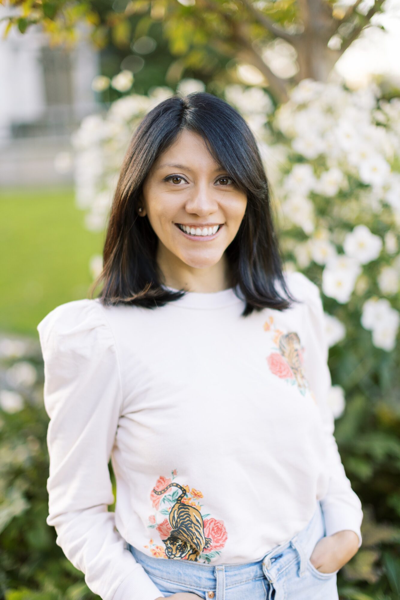 Young woman with dark hair smiling outdoors, wearing a white top with floral patterns, in front of greenery and white flowers.