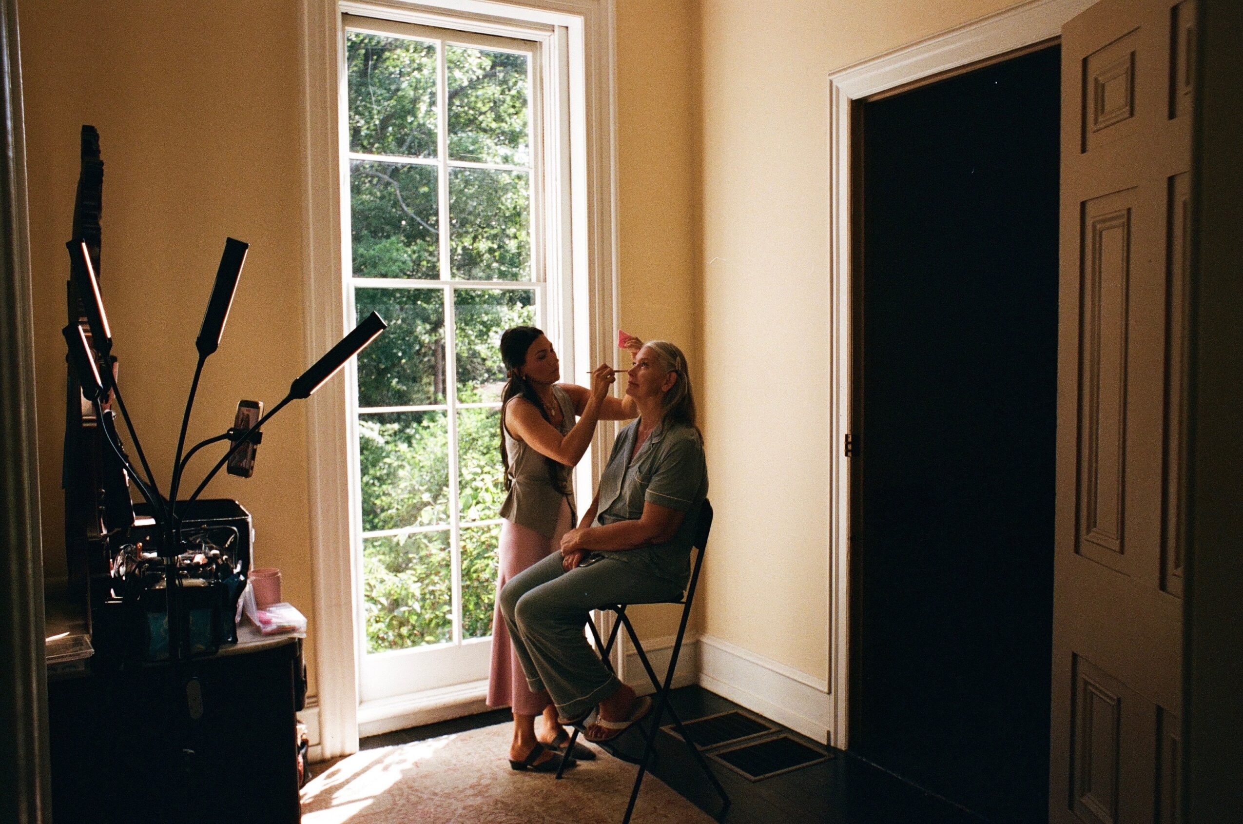 Two women in a room near a large window, one applying makeup to the other, with a door on the right.
