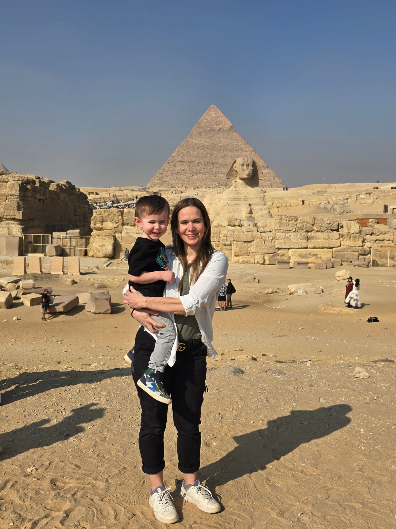 Two women stand on sandy ground with pyramids in the background under a clear blue sky.