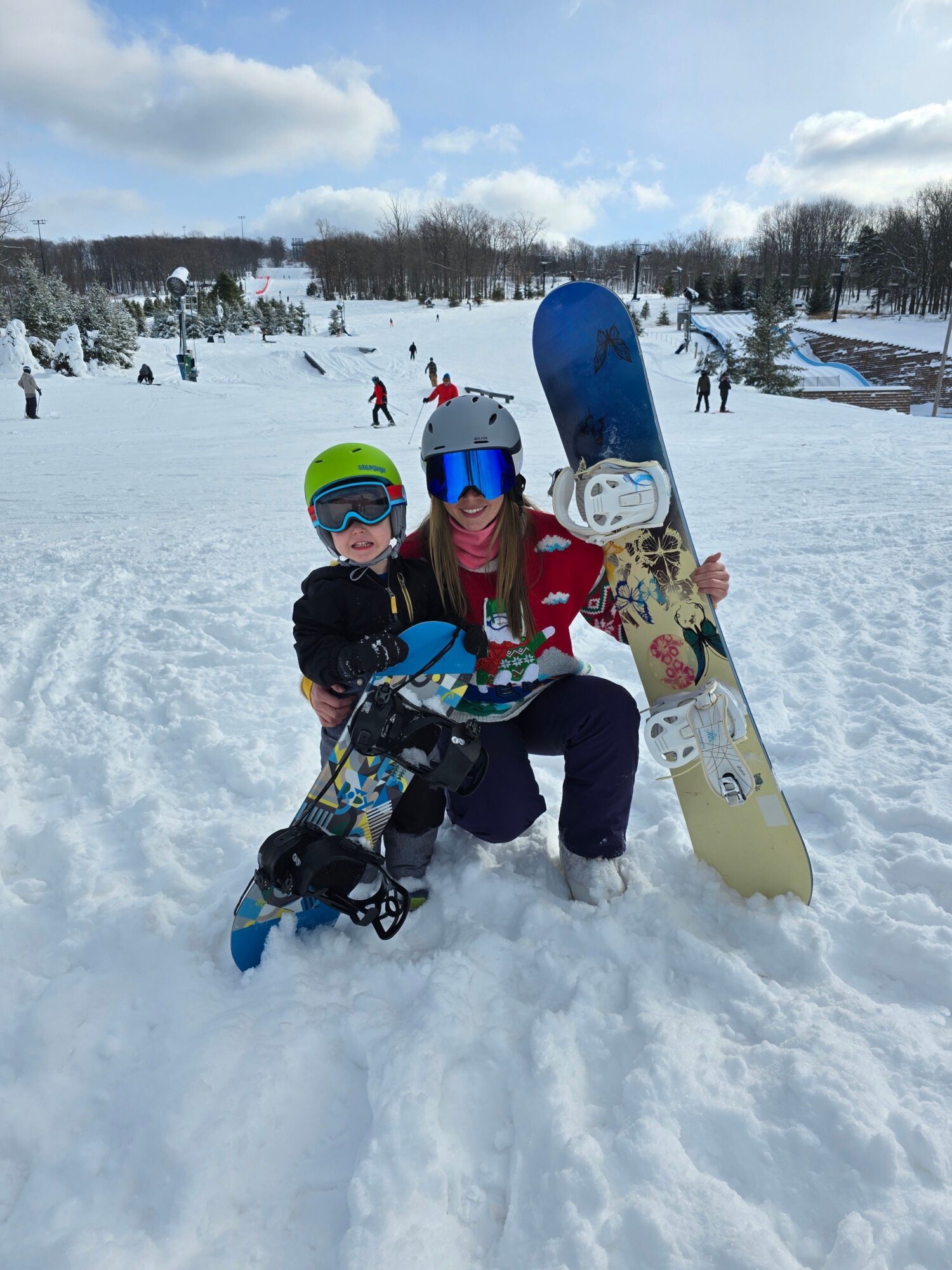 Child in winter clothing with helmet and goggles, holding a snowboard, standing in snow with trees and buildings in background.