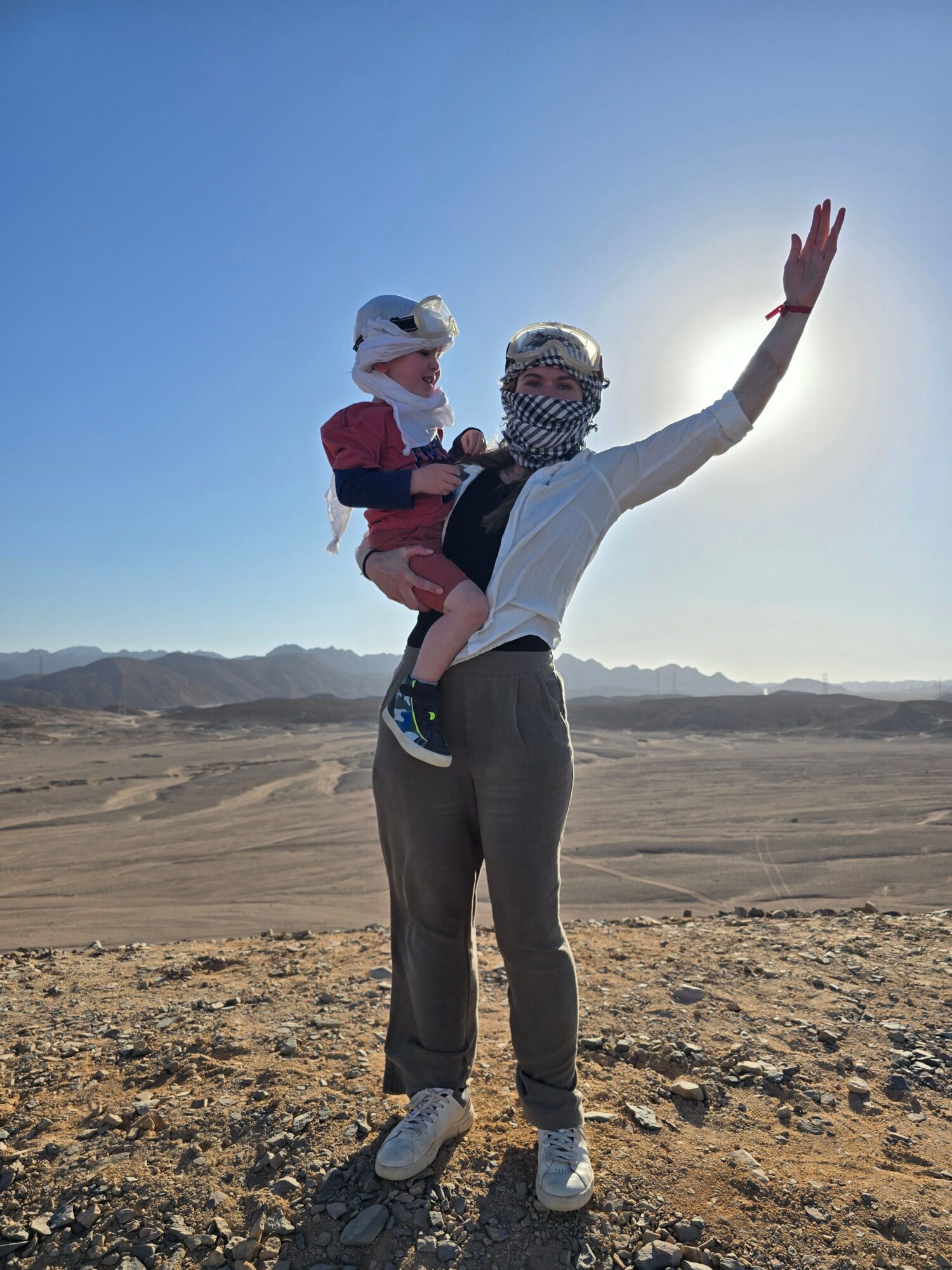 Person standing on rocky terrain with arms raised, wearing a mask, sunglasses, and casual clothing, in a desert landscape.