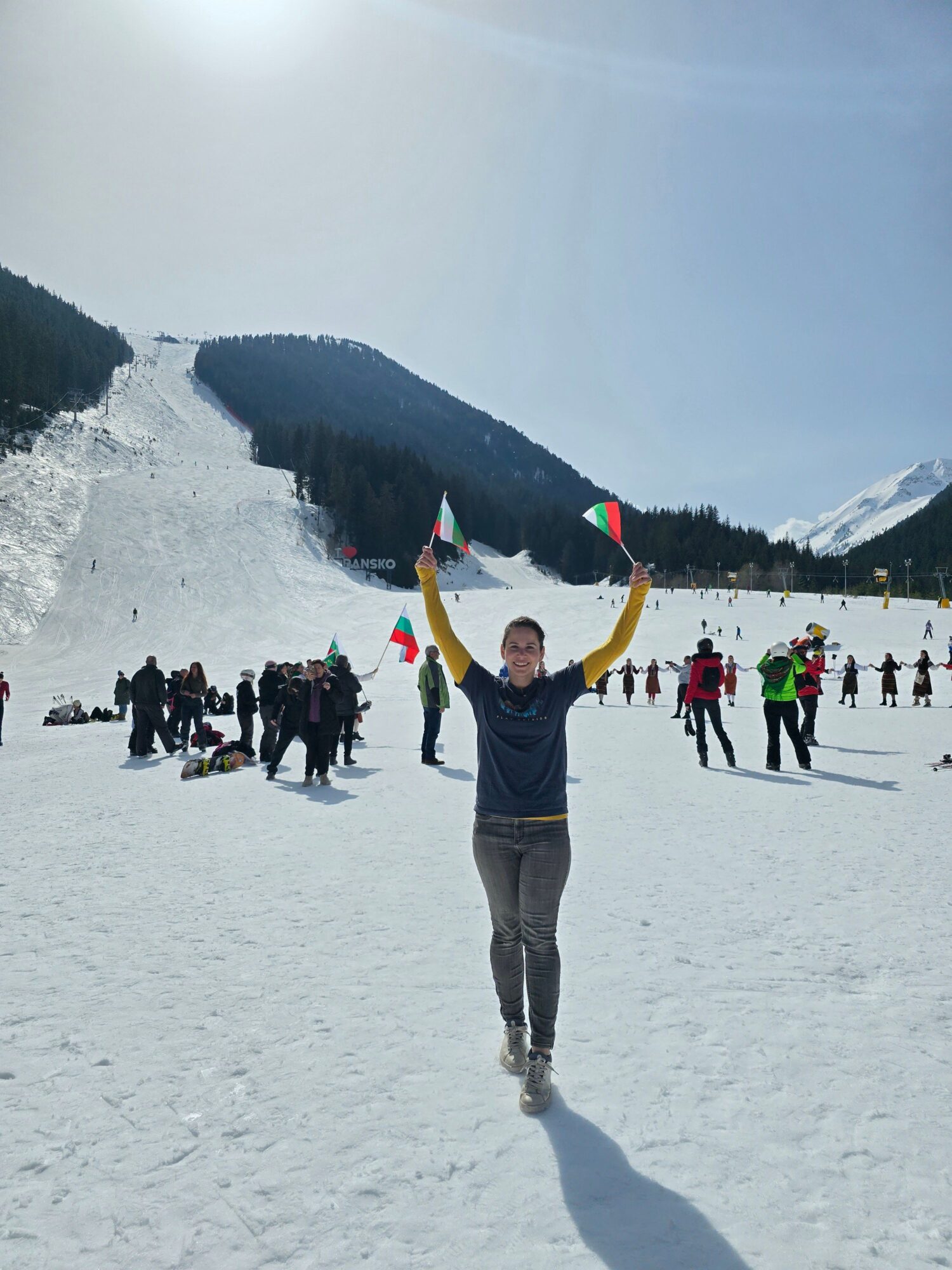 Person standing on snow with mountains and group of people in background, facing forward, in a winter landscape.