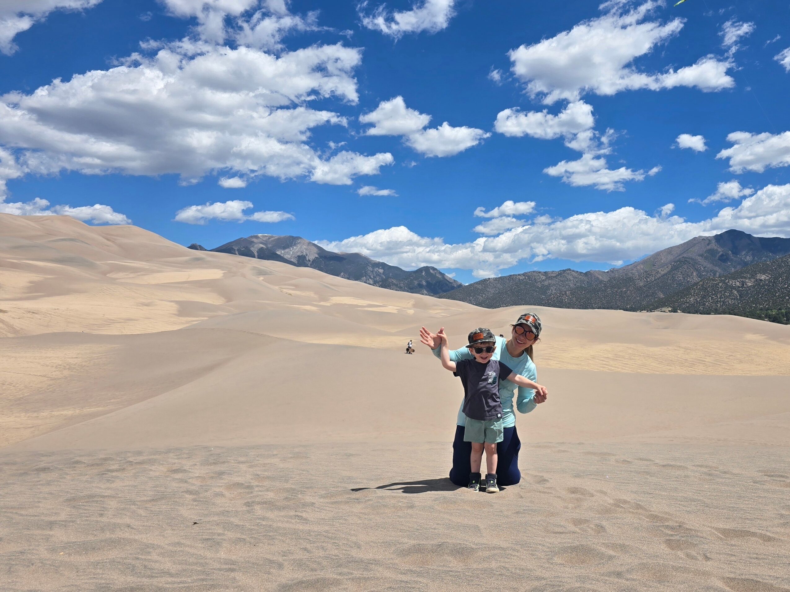 Two children stand in a desert with sand dunes, mountains, and a blue sky with clouds in the background.