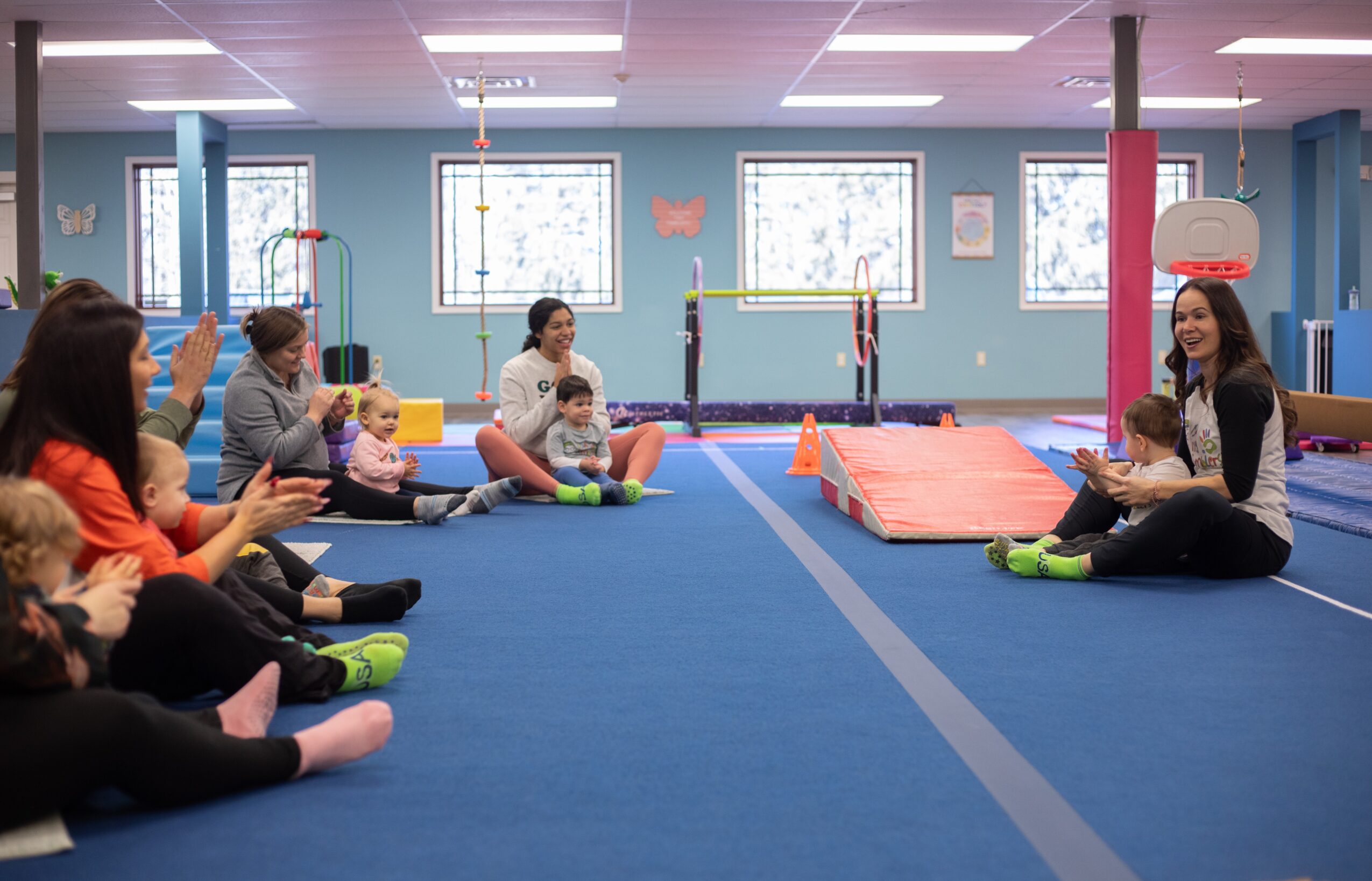Children and instructors sit on a blue gym floor in a spacious room with windows, mats, and gym equipment, engaging in a group activity.