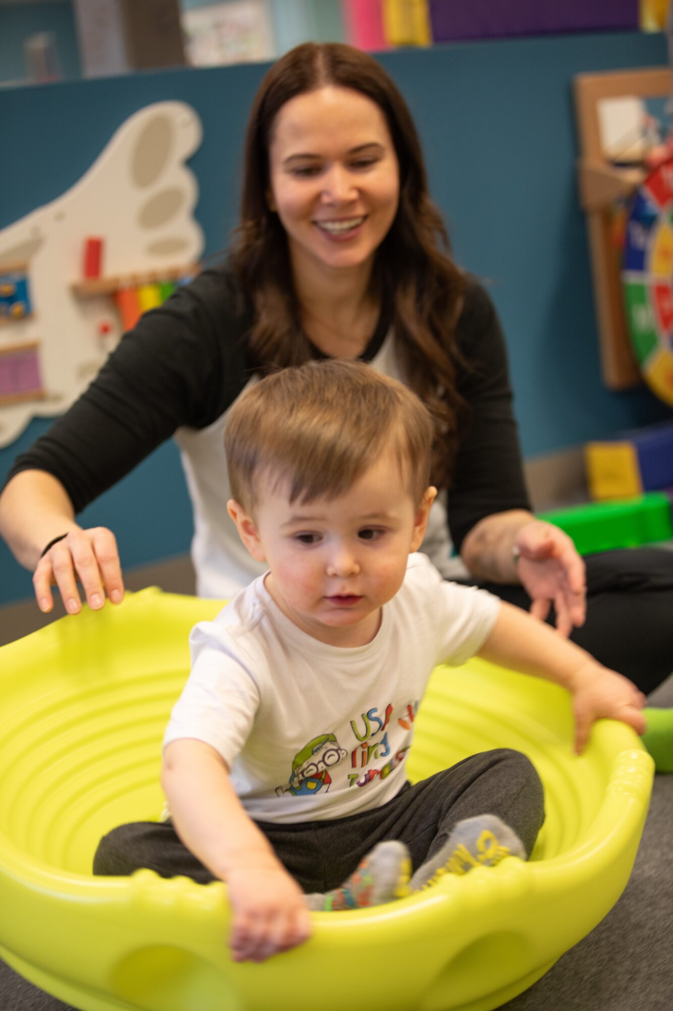 Young woman smiling behind a child sitting in a yellow plastic tub, in a colorful play area.