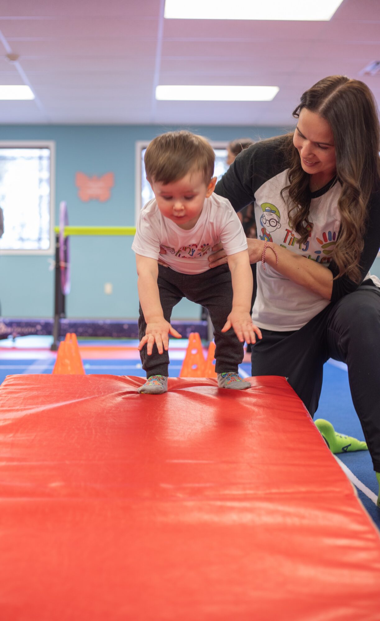Young boy and woman at an indoor play area, boy is crawling on a red mat with orange cones, woman supports him.