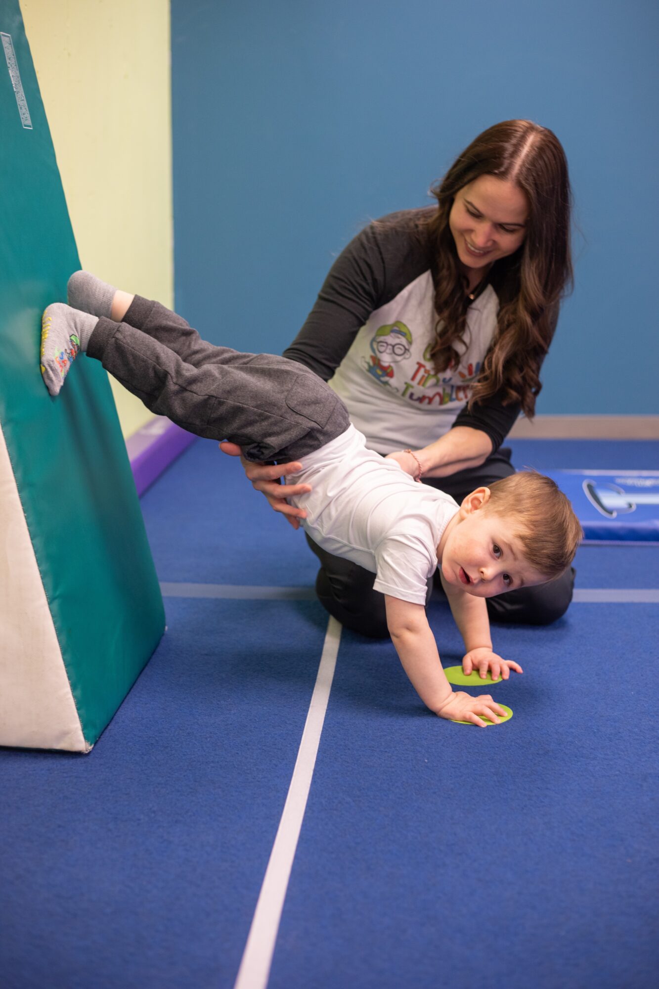 Young boy doing a handstand against a wall with a woman supporting him on a blue floor.