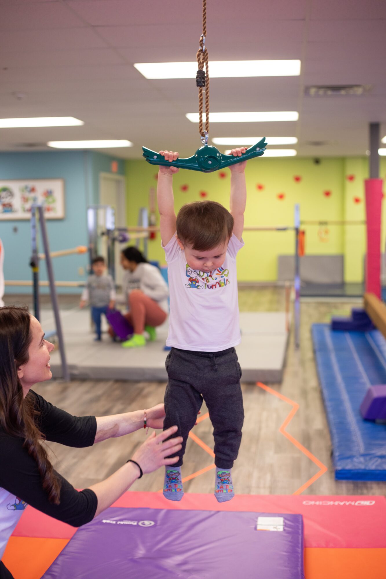 Child hanging from a trapeze in a play area, with a woman reaching up to assist, colorful mats and equipment around.