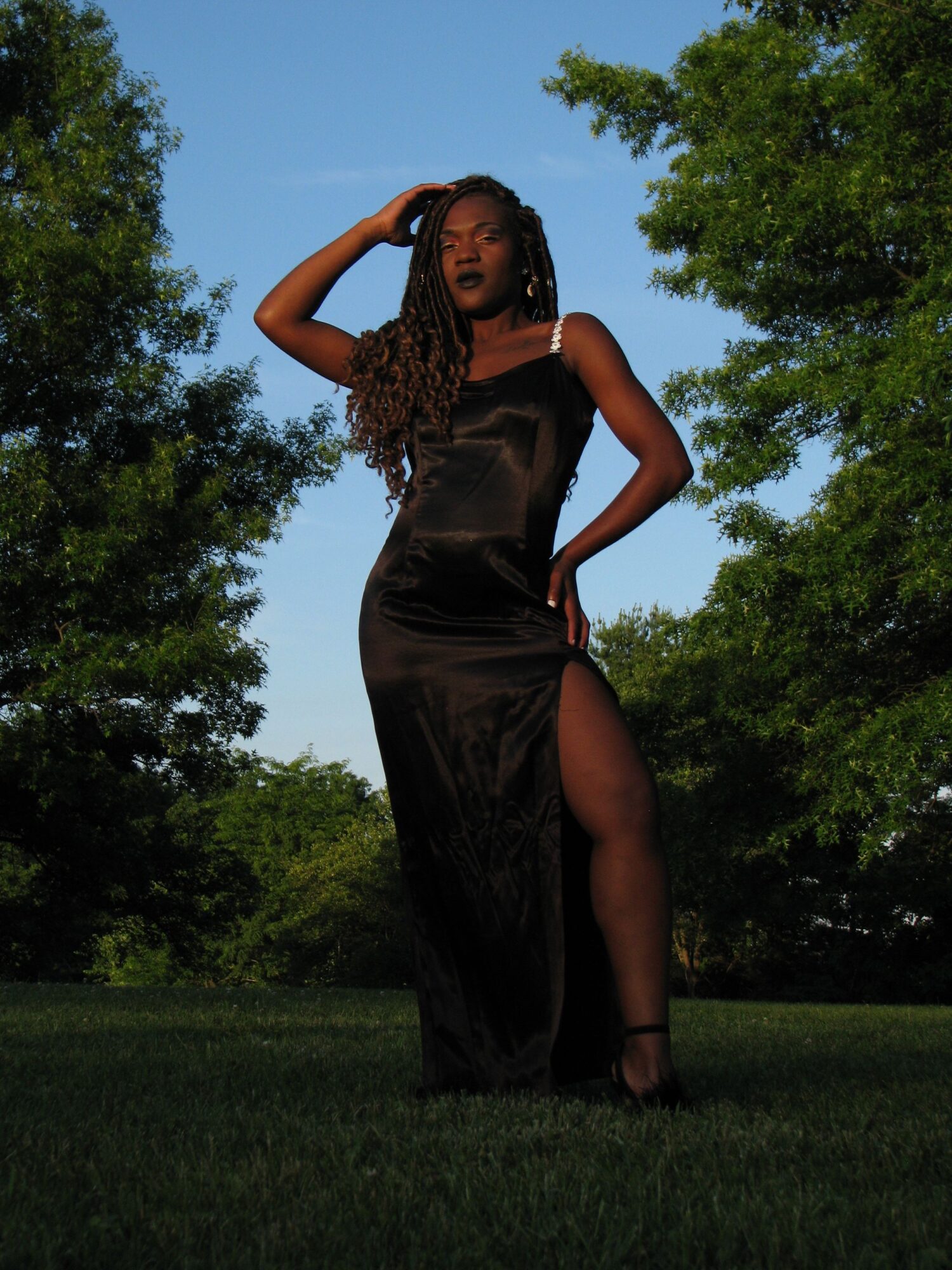 Woman in a black dress standing outdoors on grass with trees and blue sky in background.