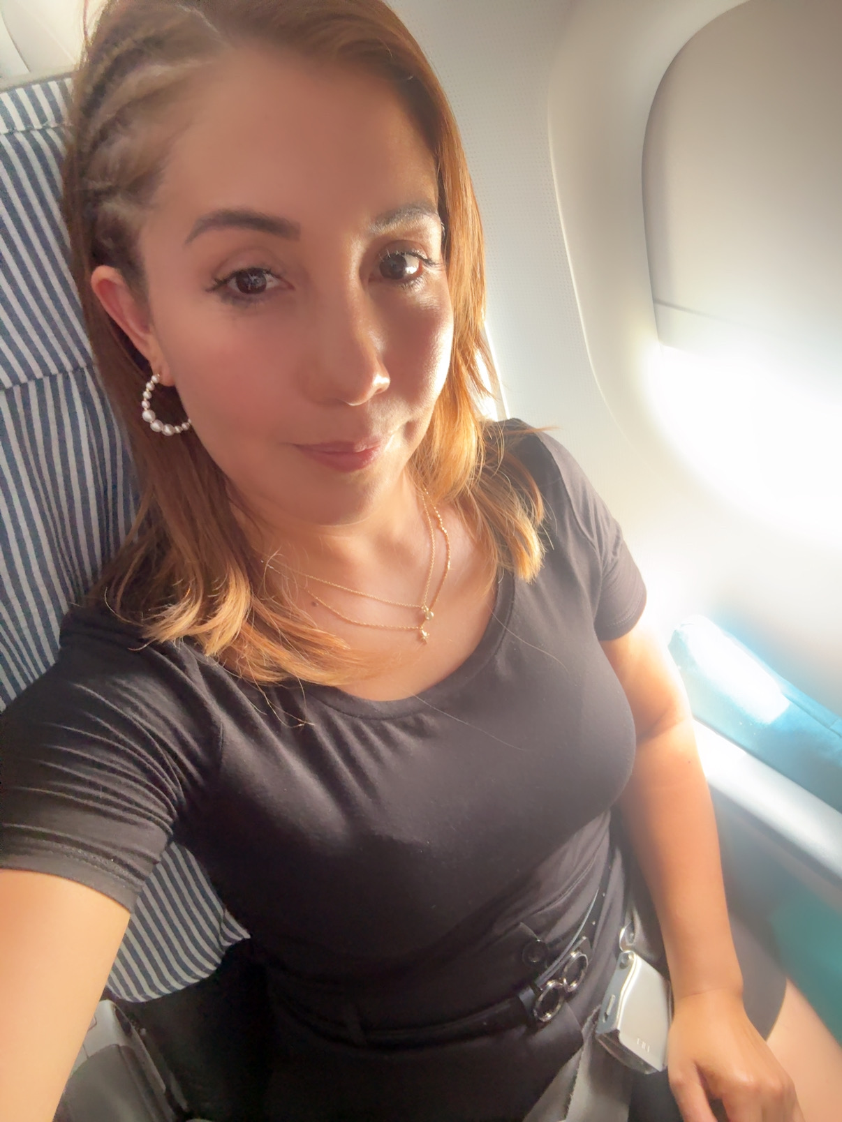 Woman with shoulder-length hair, wearing earrings and a black top, sitting by airplane window, smiling.